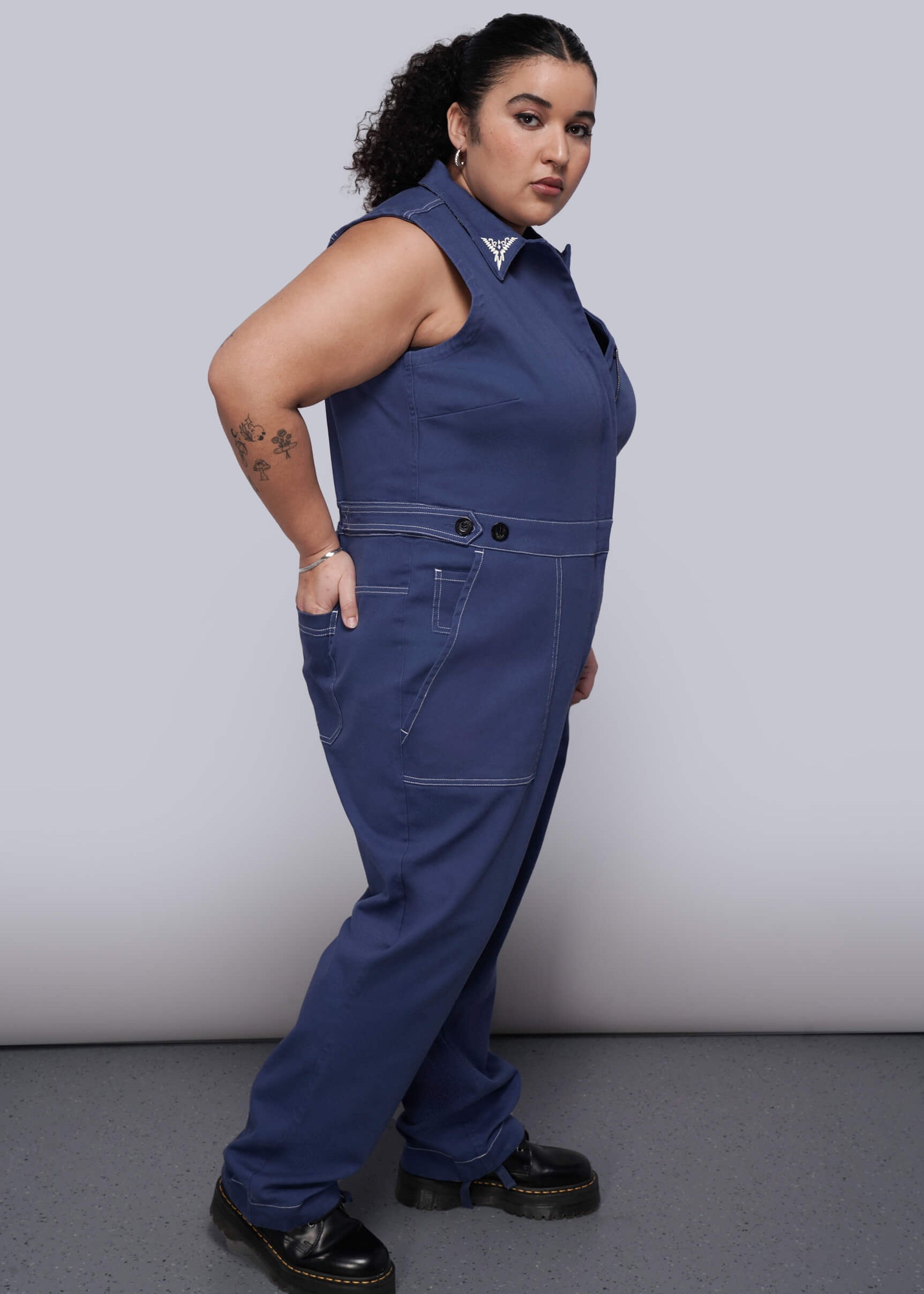 1X=Person wearing a blue sleeveless jumpsuit against a plain background