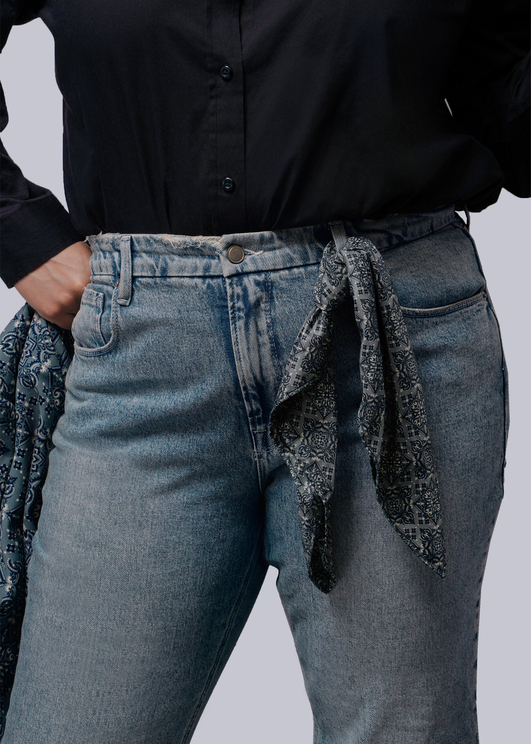 Person wearing a black shirt and blue jeans with a patterned scarf tied around their waist on a plain background