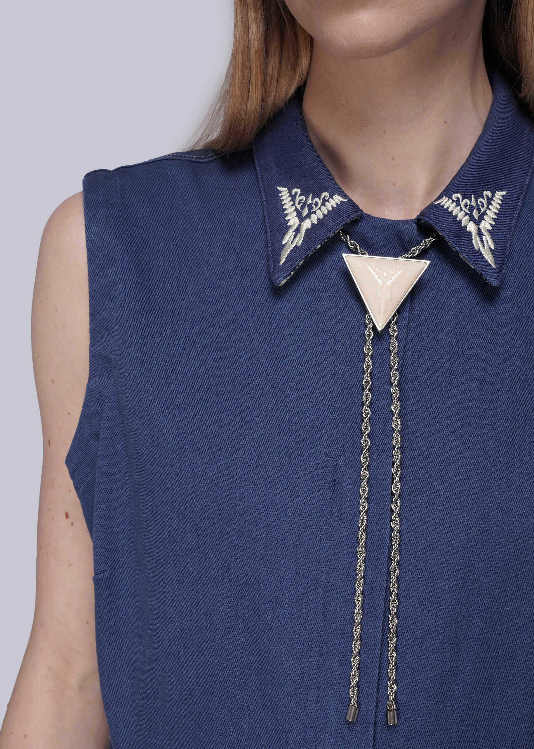 Close up of woman wearing blue denim coverall with white embroidered collar and silver chain bolo tie with resin pendant.
