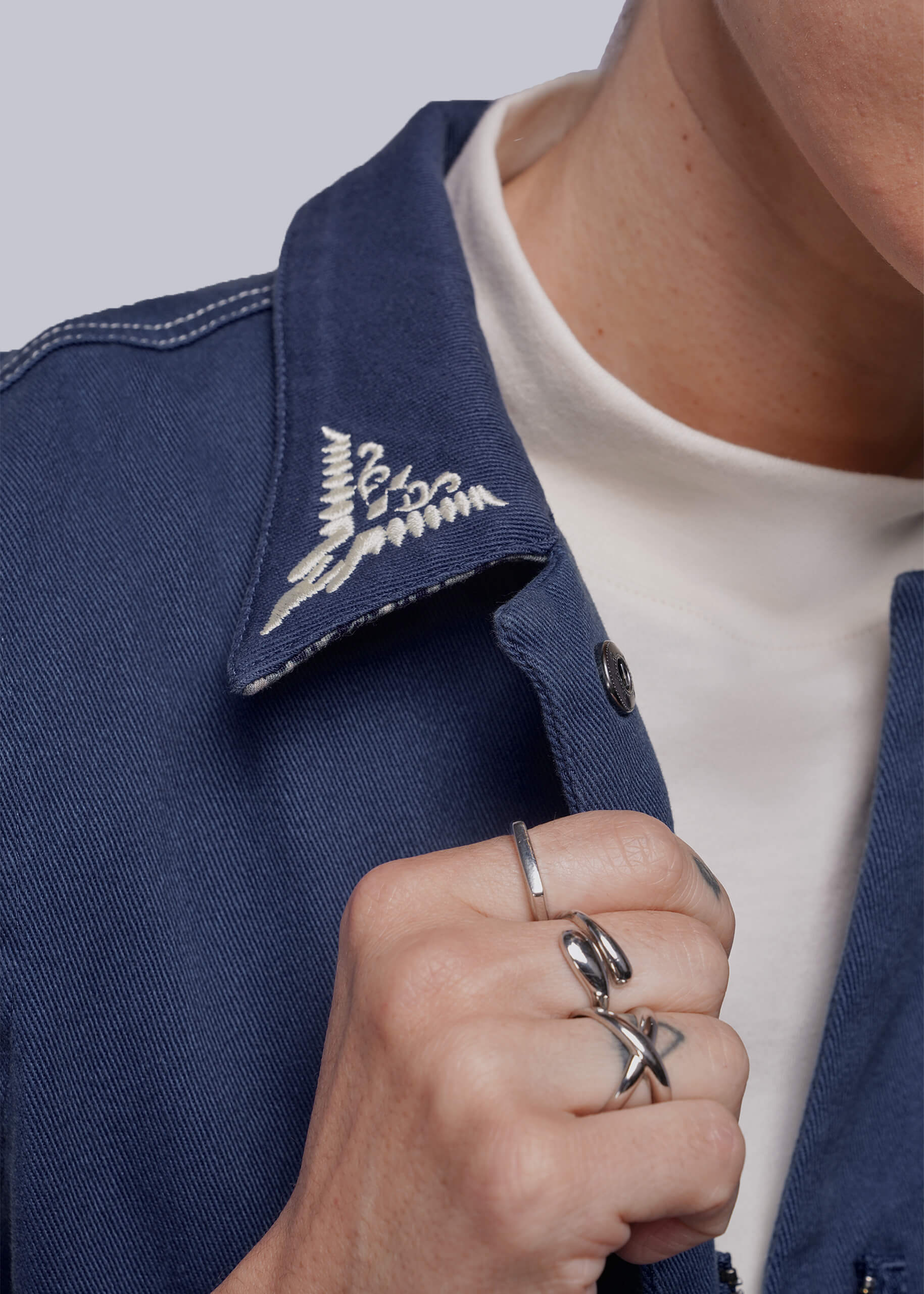 S=Close-up of a person wearing a blue jumpsuit with collar embroidery, a white shirt, and multiple rings.