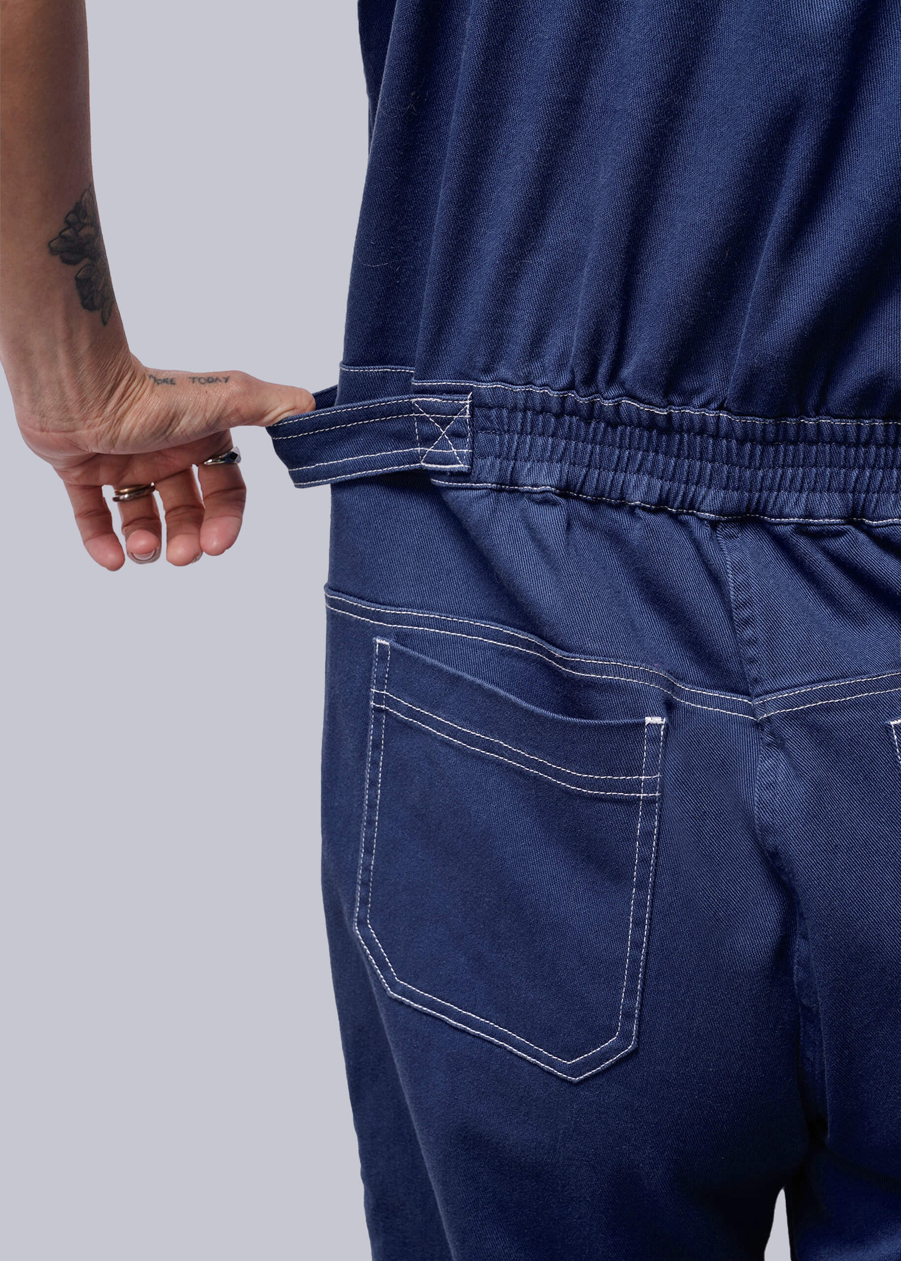 S=Close up of person wearing a navy blue jumpsuit holding elastic waistband with a plain background