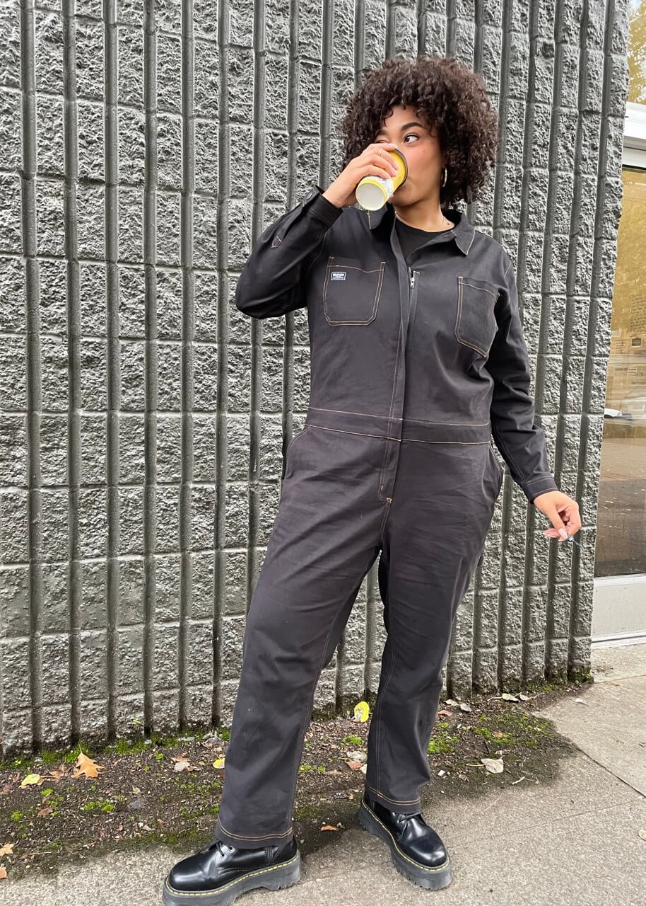 1X=Wearing The Essential Long Sleeve Coverall in an oversized fit, a person with short curly hair sips from a cup, standing against a textured gray wall. They sport black shoes while scattered leaves on the ground enhance the cozy autumn vibe.