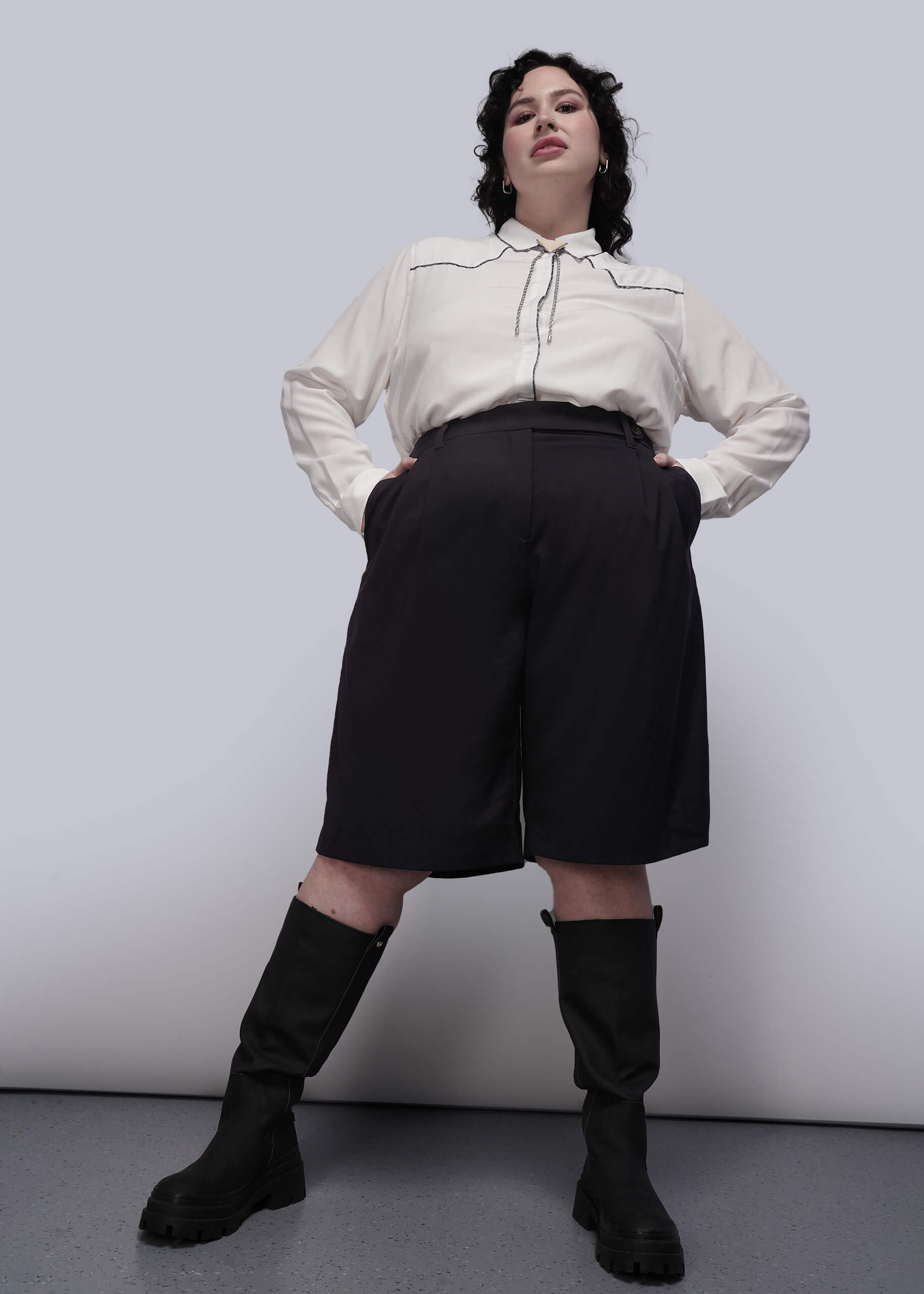 1X=Person wearing a white blouse, black high-waisted pants, and black knee-high boots against a plain background