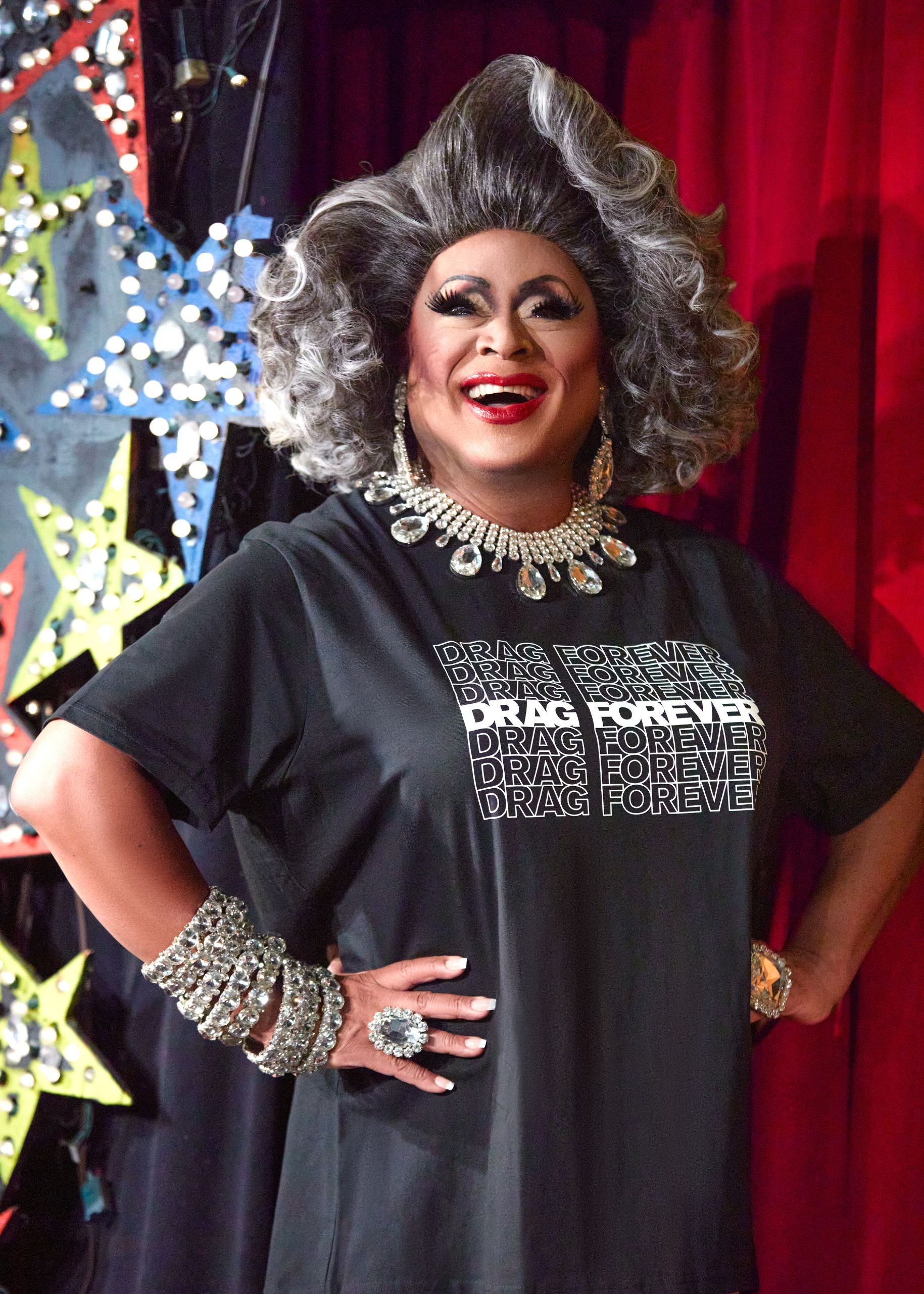 Person wearing a black t-shirt with 'Drag Forever' text, posing in front of a colorful and vibrant background.
