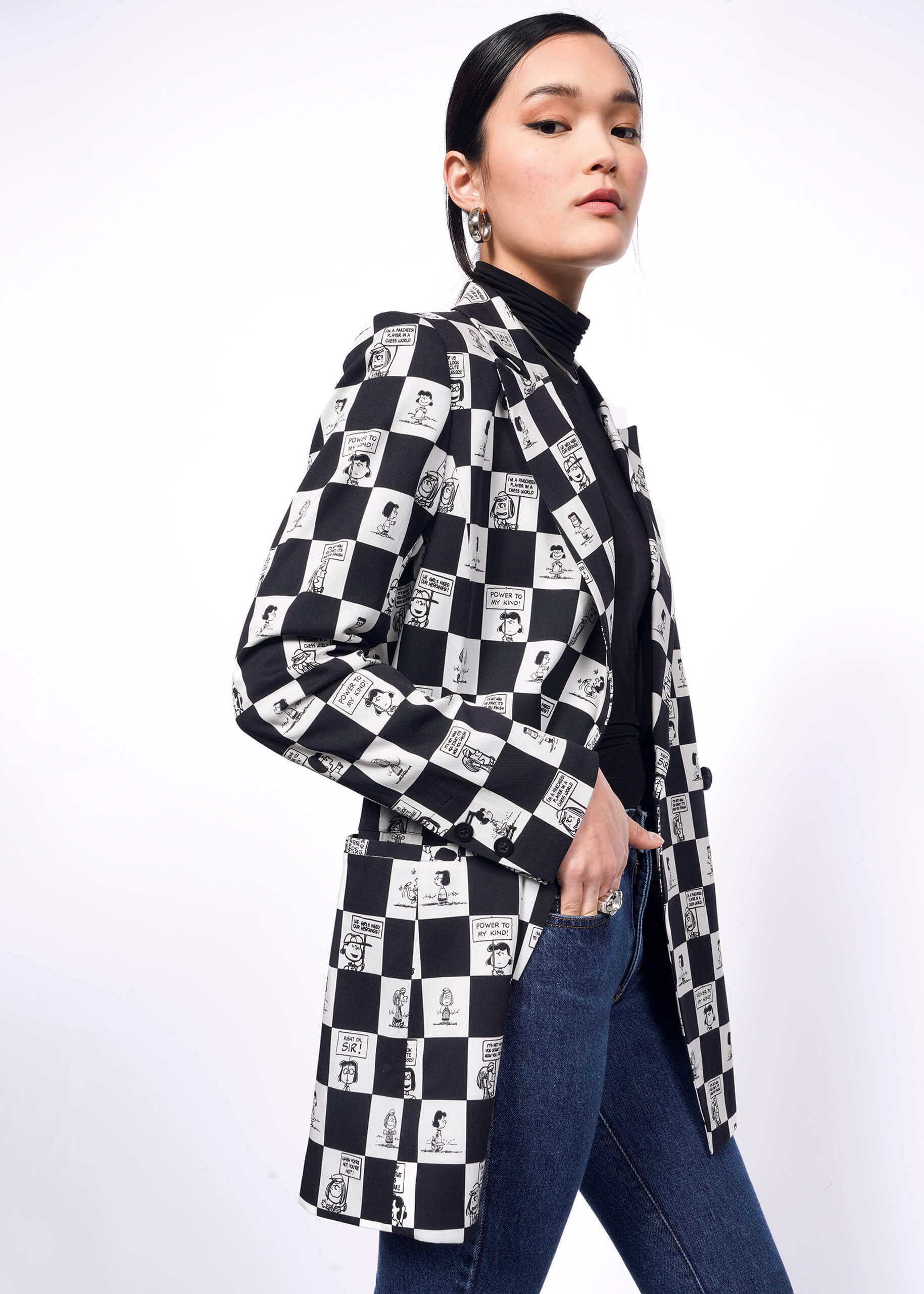 S=Side view of model with black hair tied back wearing a black turtleneck, jeans, and The Peanuts x Wildfang Double Breasted Blazer in a custom black and white checkered print featuring Peanuts characters.