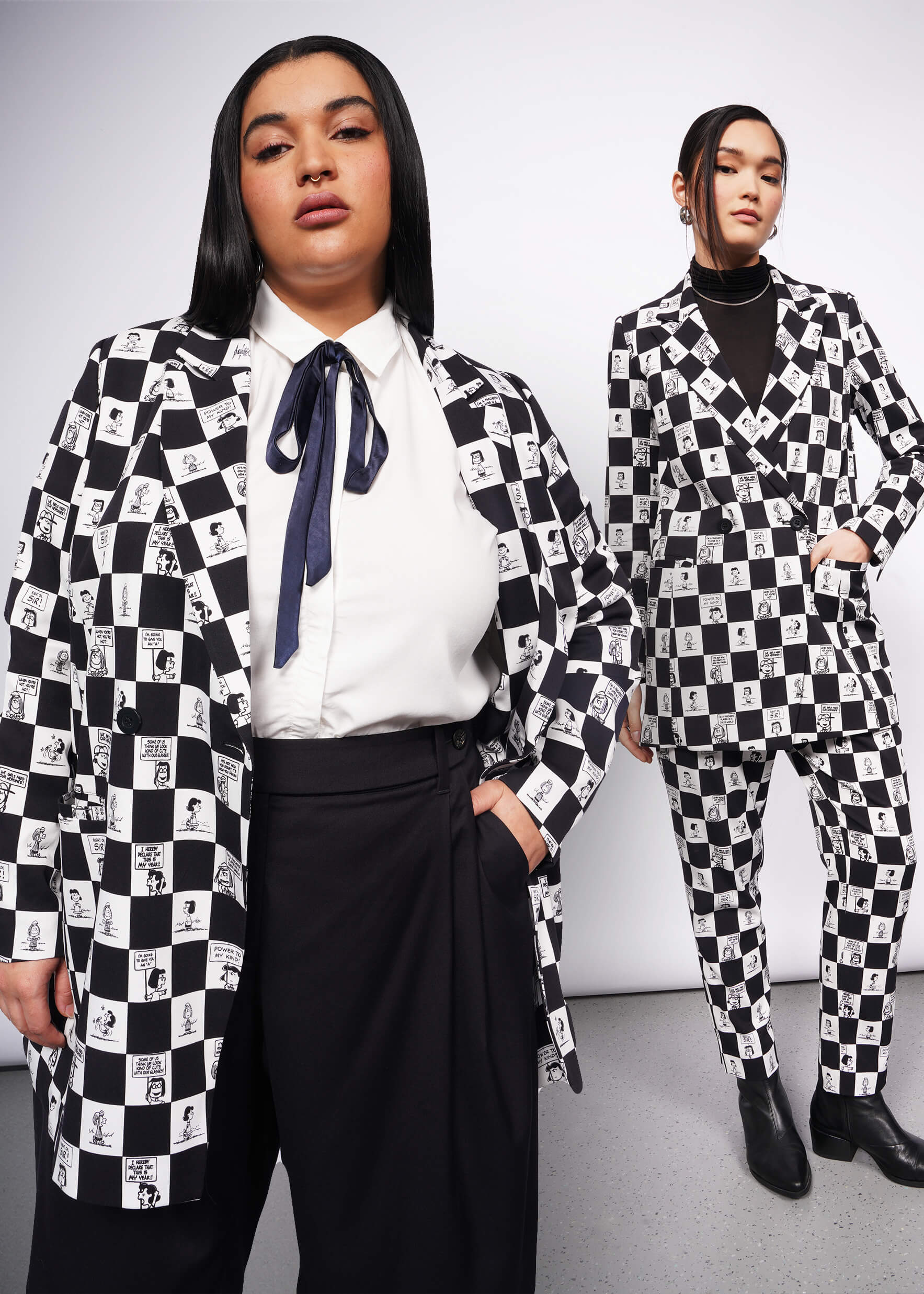 Two models wearing The Peanuts x Wildfang Double Breasted Blazer in a custom black and white checkered print featuring Peanuts characters. One model wears the matching Slim Crop Pant, and the other styles it with The Empower bOW Button Up and a black trouser.