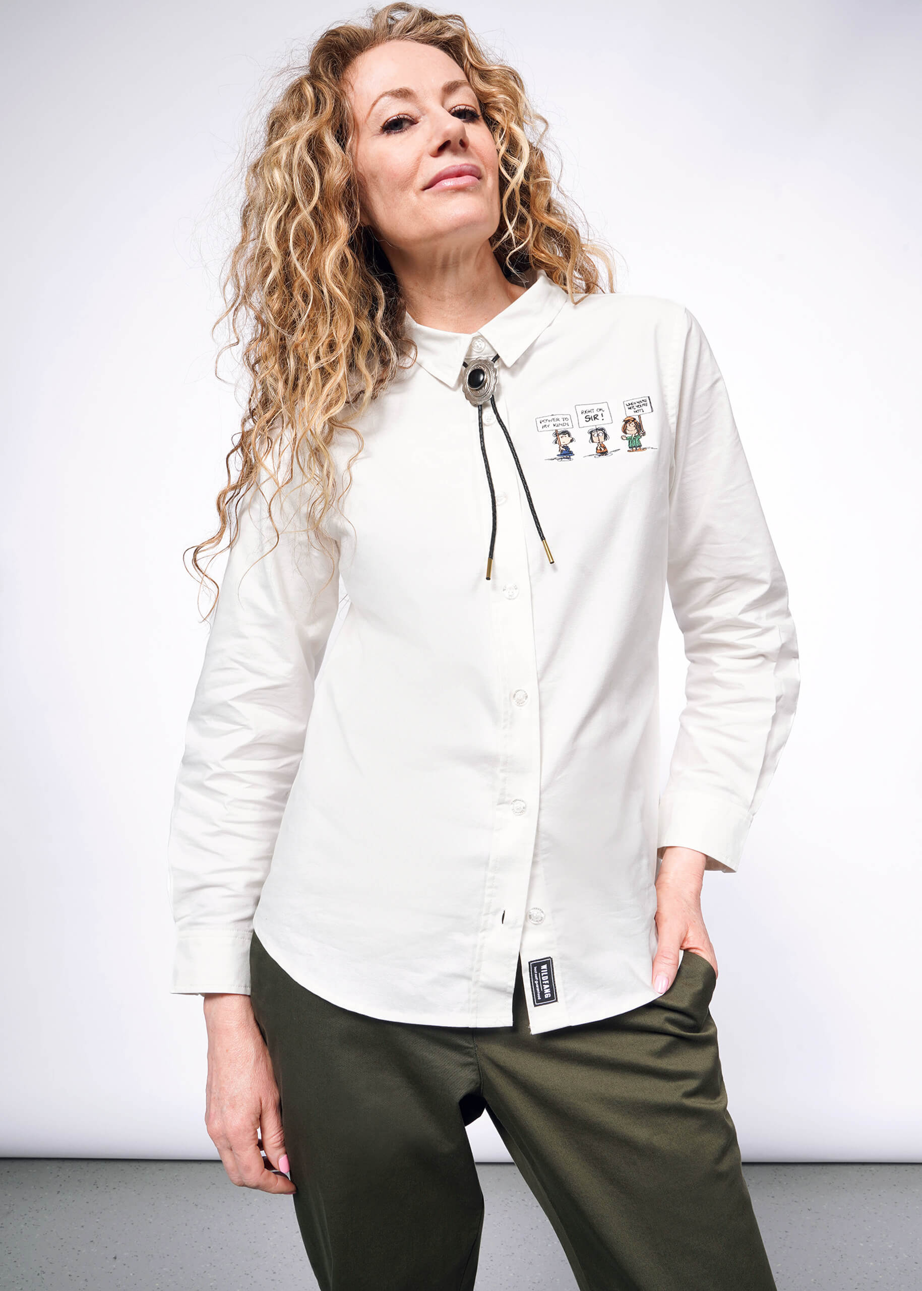 S= Person with wavy blonde hair looking at camera wearing The Peanuts x Wildfang Long Sleeve Button Up, which is a white long sleeve button up shirt with embroidered Peanuts characters, green trousers, and bolo tie.