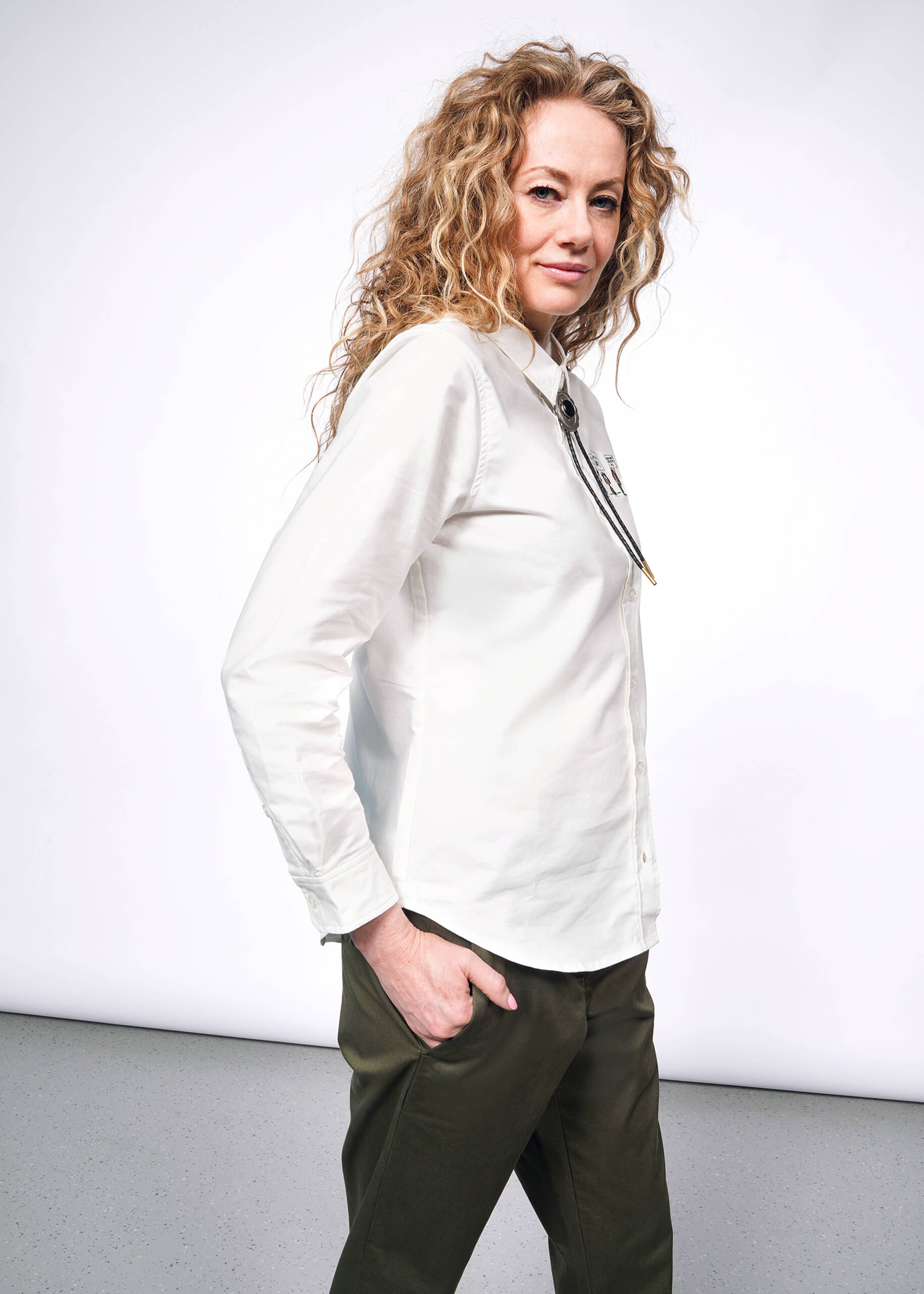 S=Side view of person with wavy blonde hair looking at camera wearing The Peanuts x Wildfang Long Sleeve Button Up, which is a white long sleeve button up shirt with embroidered Peanuts characters, green trousers, and bolo tie.