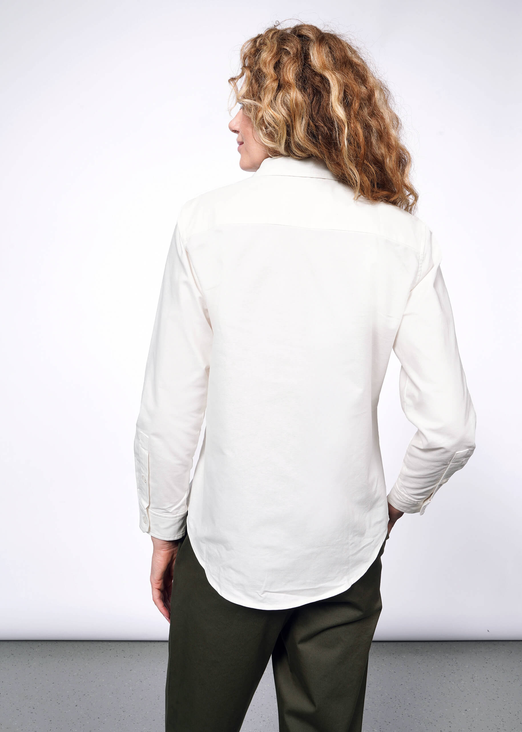 S= Back view of person with wavy blonde hair looking at camera wearing The Peanuts x Wildfang Long Sleeve Button Up, which is a white long sleeve button up shirt with embroidered Peanuts characters (embroidery not in view here), and green trousers.