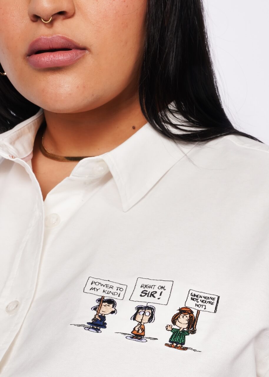 1X=Close up of person wearing The Peanuts x Wildfang Long Sleeve Button Up, which is a white button up shirt with the detail of embroidered Peanuts characters on left chest.