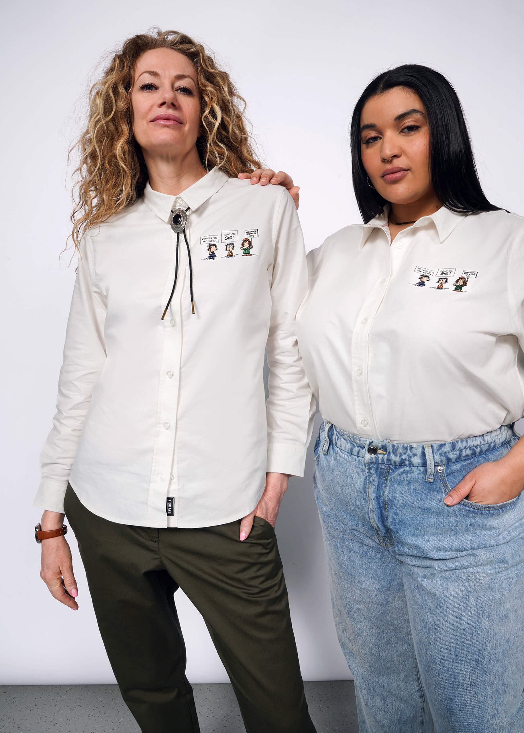 Two models stand side by side wearing The Peanuts x Wildfang Long Sleeve Button Up, which is a long sleeve button up with Peanuts character embroidery on the left chest.