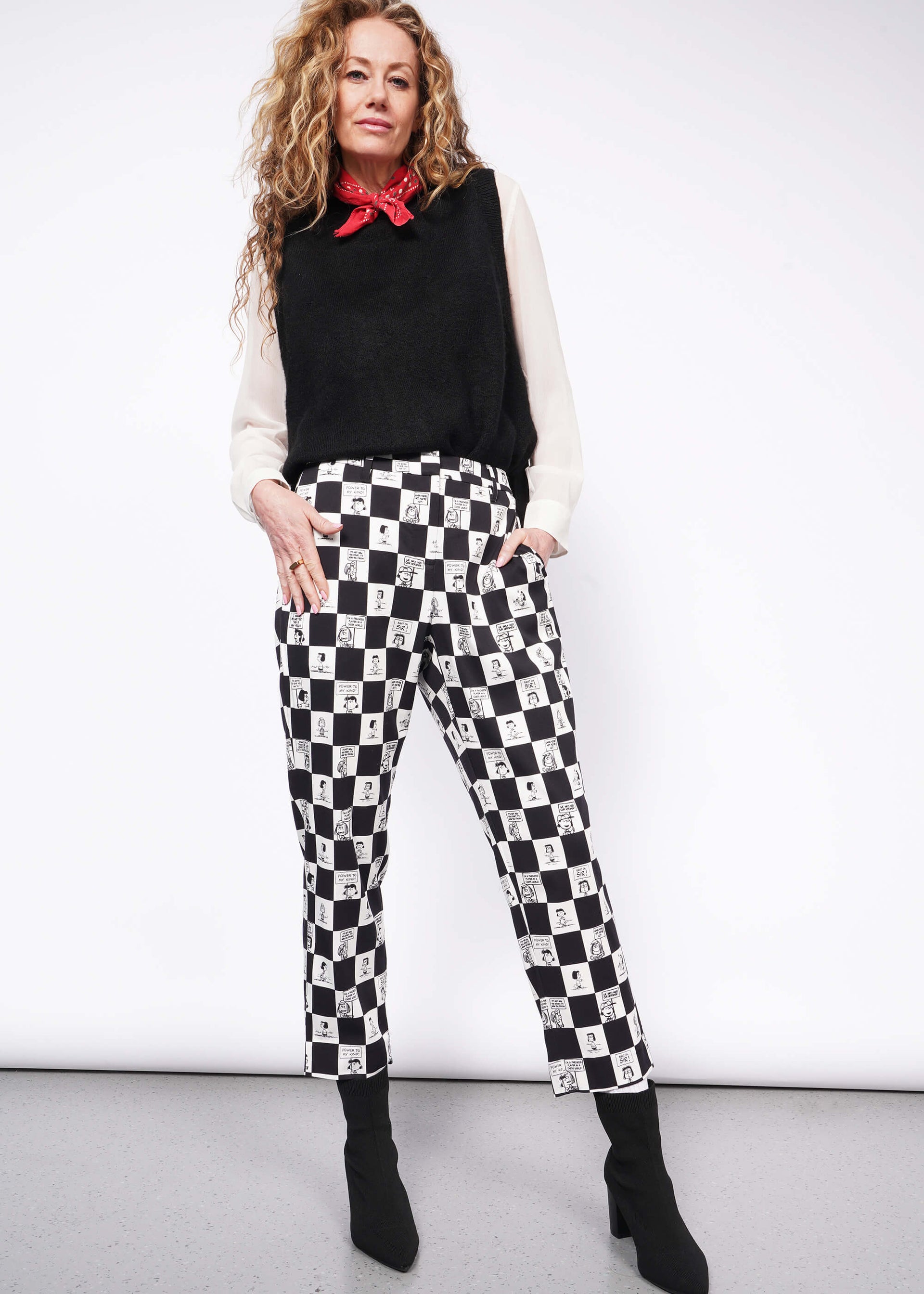 S= Person with wavy blonde hair looks at camera wearing white long sleeve button up under black sweater vest, a red bandana tied around their neck, black heeled boots, and the checkered black and white Slim Crop Pant from the Peanuts x Wildfang collection.