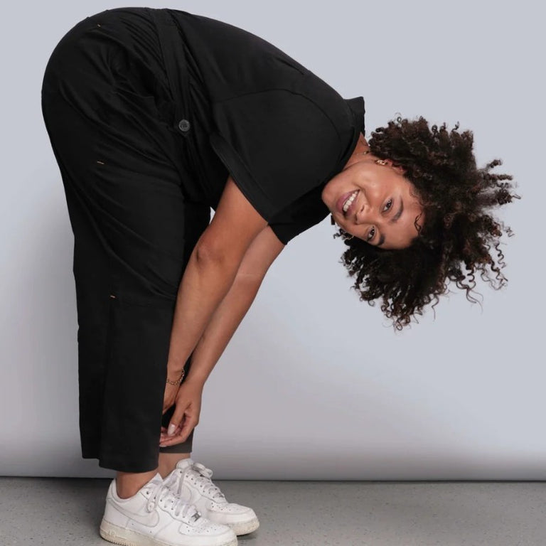 Model wearing black coverall showing stretch by bending at waist to touch toes.