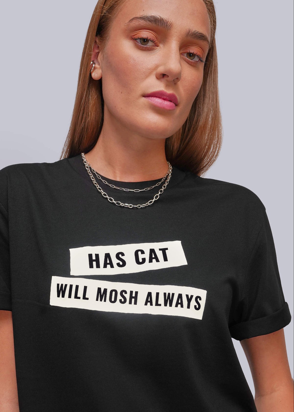 S=A woman wearing a black short-sleeve graphic t-shirt with white rectangular blocks containing the text 'HAS CAT' and 'WILL MOSH ALWAYS' in black capital letters. The background is a solid light gray studio backdrop.