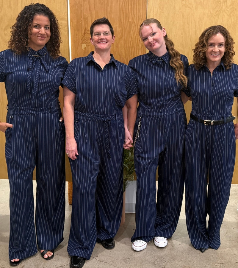 Image of four people wearing the same navy blue pinstriped wide leg jumpsuit standing in front of light wood paneled wall.