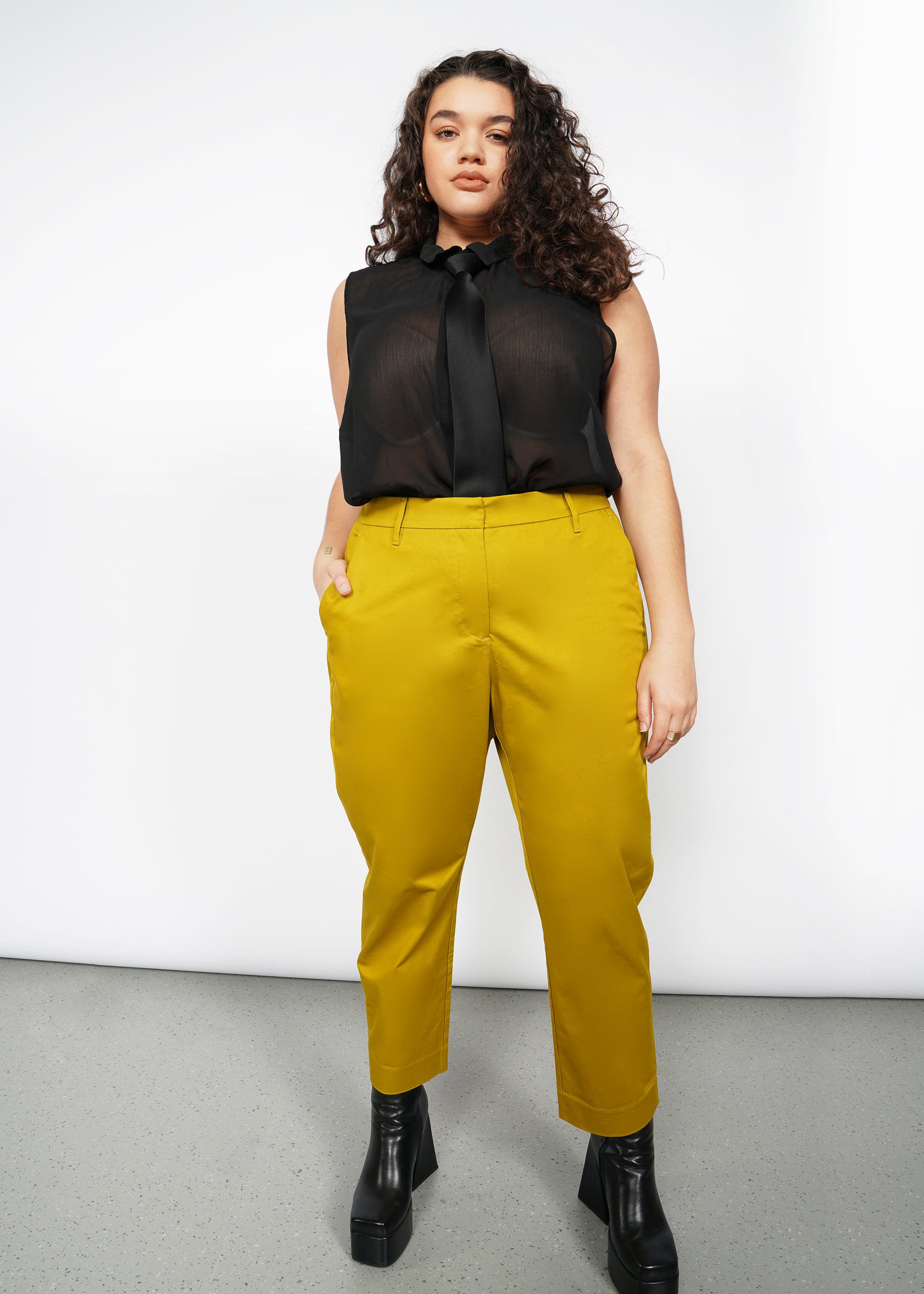 1X=A woman with long curly hair wears a sheer black sleeveless blouse paired with The Essential Slim Crop Pant in mustard. She completes her look with black platform boots, standing confidently against a plain white background, her hands in her pockets.
