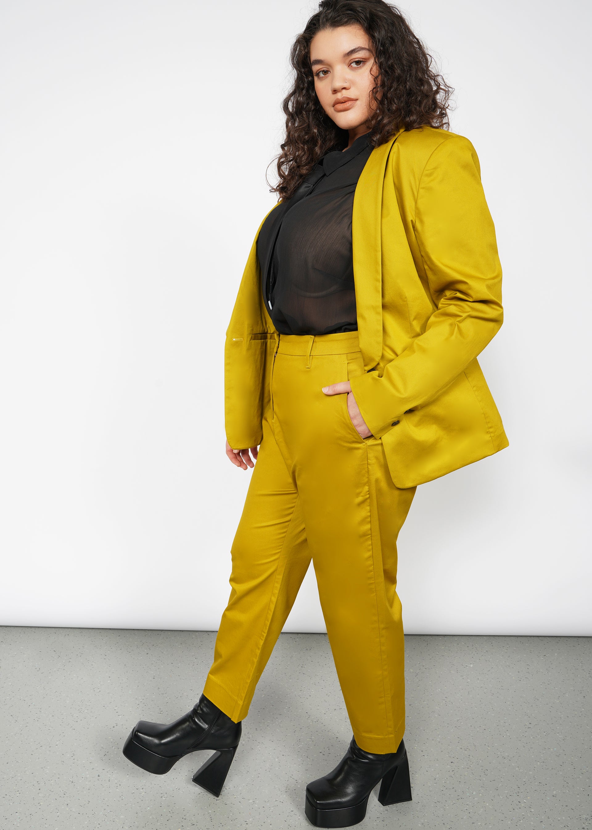 1X=A person with long curly hair confidently wears a bright yellow suit featuring The Essential Slim Crop Pant, offering a breathable tailored mid-rise fit, paired with a sheer black blouse. They have black platform shoes and pose against a plain white background.