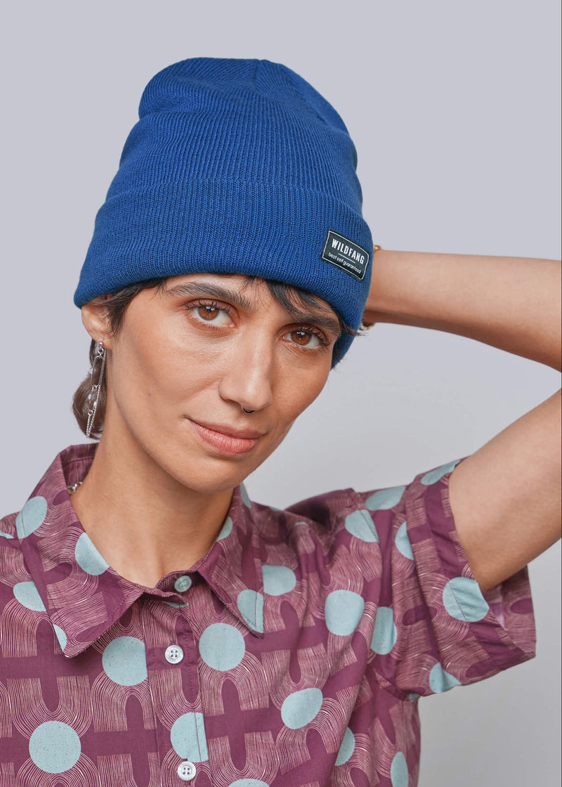 A person wearing a blue ribbed knit beanie with a small black rectangular brand patch that reads 'WILDFANG' and a mauve short-sleeve button-up shirt with a light blue and mauve geometric pattern, against a plain light gray studio background.
