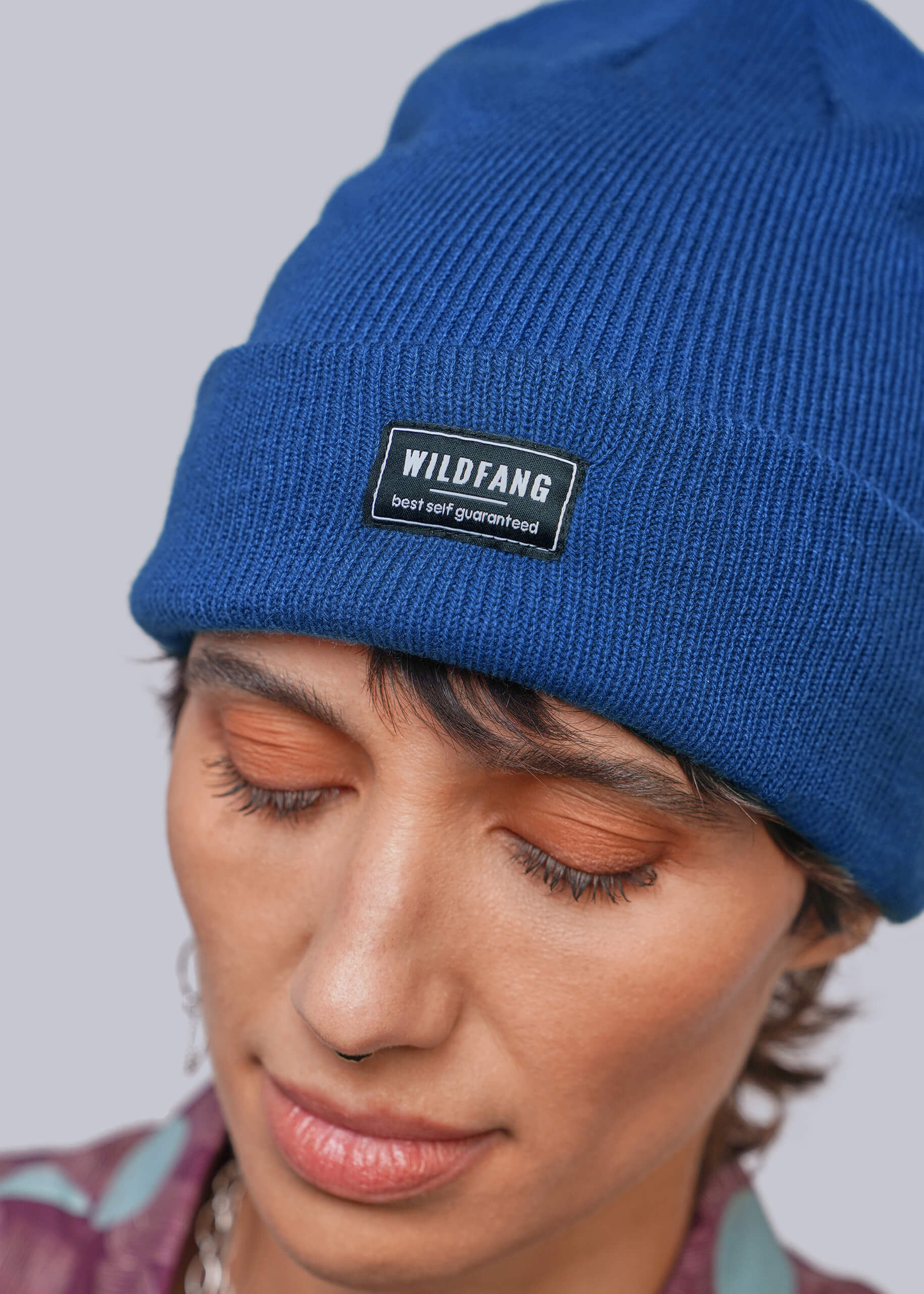 A close-up of a person wearing a royal blue ribbed knit beanie with a black rectangular patch that reads 'WILDFANG best self guaranteed.' The person is shown from the shoulders up against a light gray studio background.