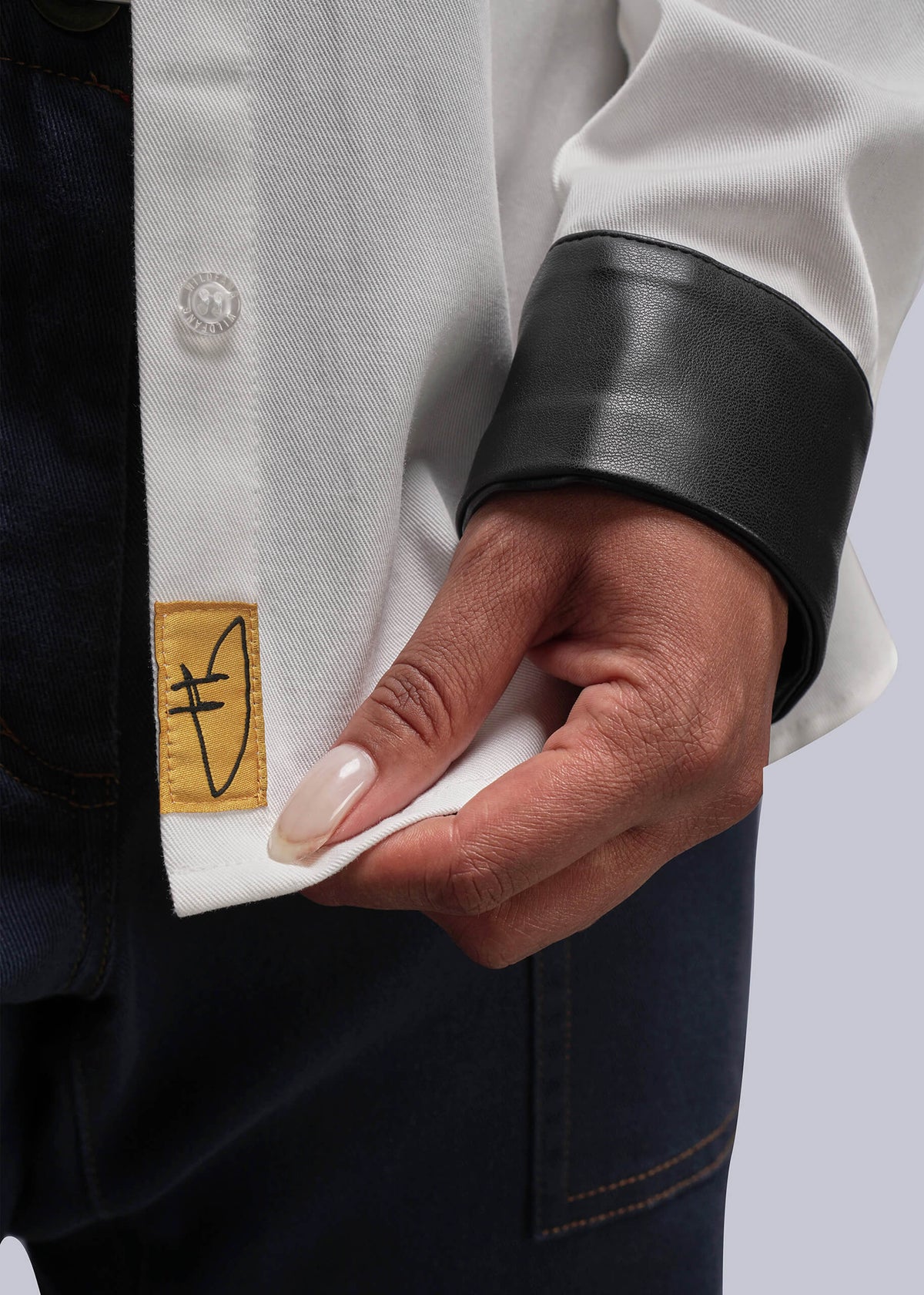 1X=Close-up of a person wearing a white button up with a black cuff and a gold brand patch on a white background