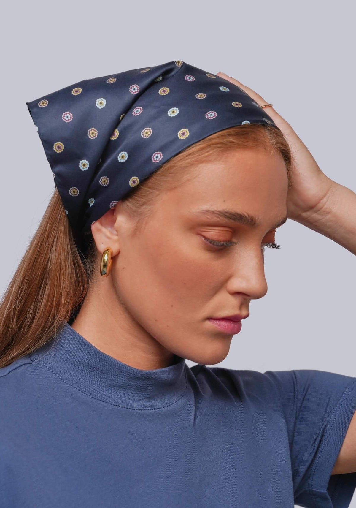 A woman wearing a navy blue silk-like headscarf with a repeating circular pattern in beige, white, and muted red, paired with a blue t-shirt against a solid gray background.