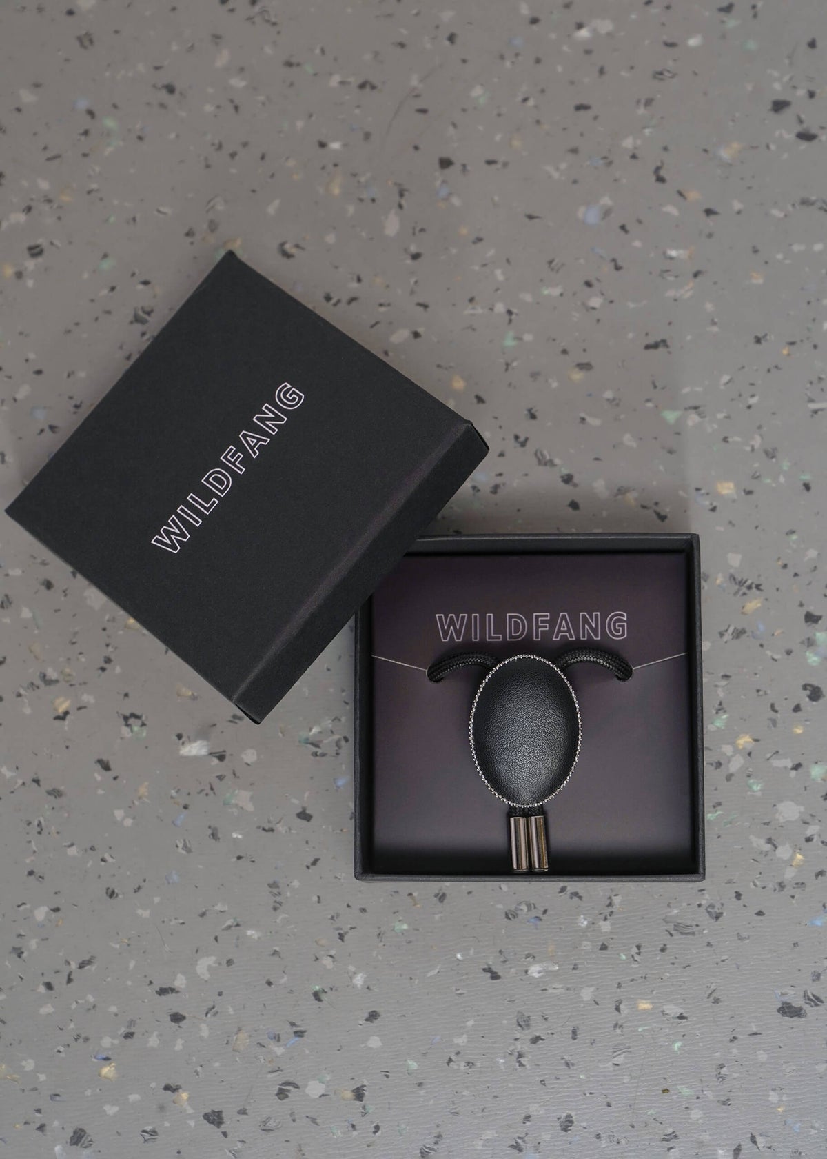A black Wildfang brand jewelry box, shown open with its lid leaning against it, revealing a black bolo tie with circular pendant matching dark display insert, all placed on a speckled gray surface.