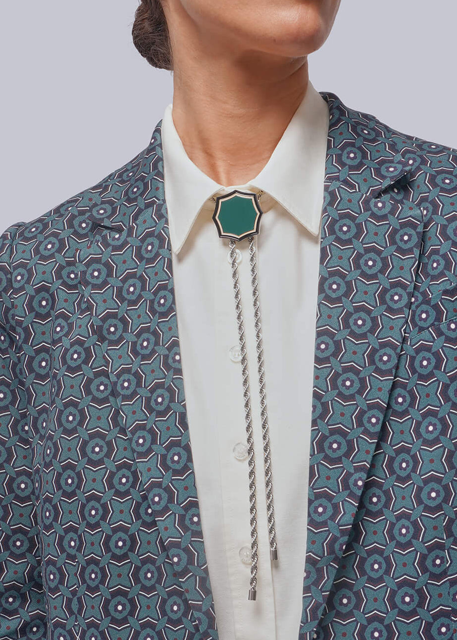 A model wearing a navy blue patterned blazer over a white collared shirt with a silver-toned bolo tie and a green pendant, set against a solid light gray background.