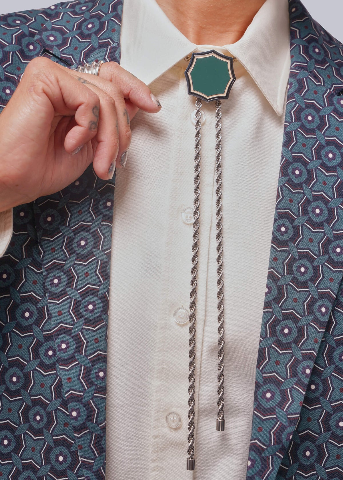 A close-up of a person wearing a silver-toned bolo tie with a green hexagonal center, styled over a white collared shirt and a blue patterned blazer.