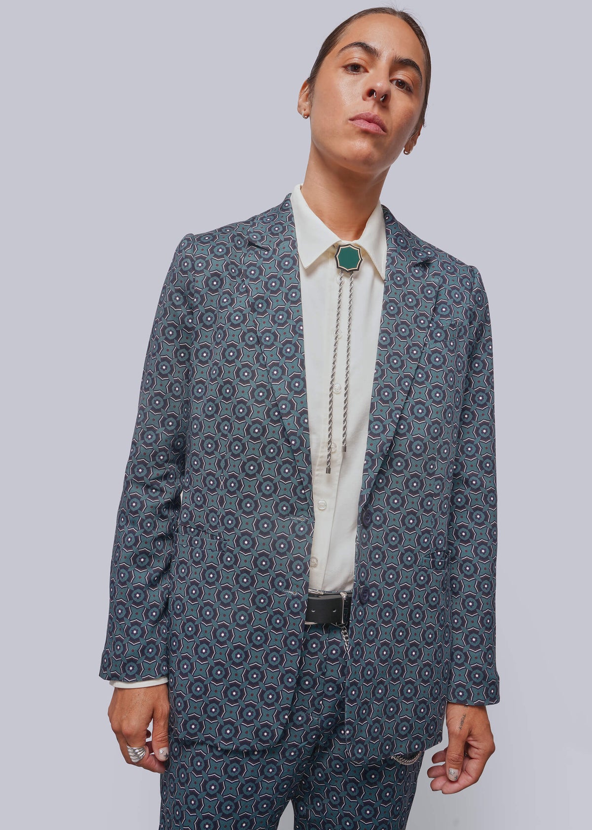 A model wearing a blue and white patterned blazer and matching trousers, styled with a white shirt, a long silver bolo tie with a green pendant, and a black belt, against a solid gray background.