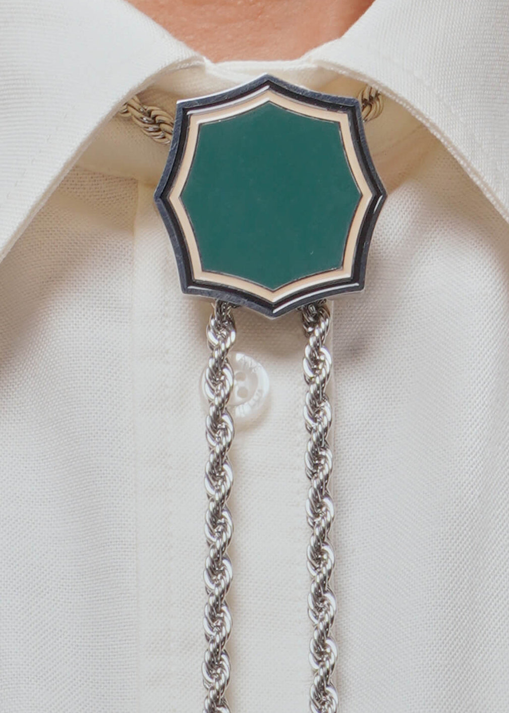 A close-up of a person wearing a silver-toned bolo tie with a green shield-shaped face and a white collared shirt, with a patterned jacket visible at the sides.