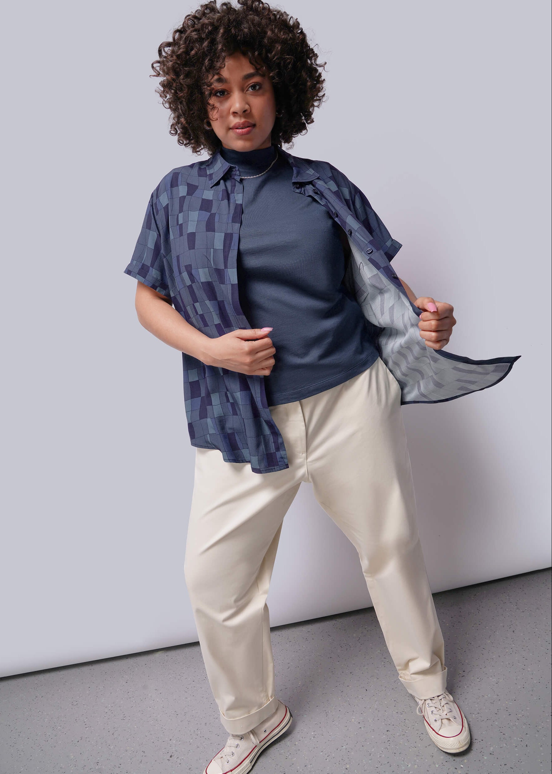1X=Person wearing a blue shirt with a patterned vest over a dark turtleneck, beige pants, and white sneakers against a plain background.