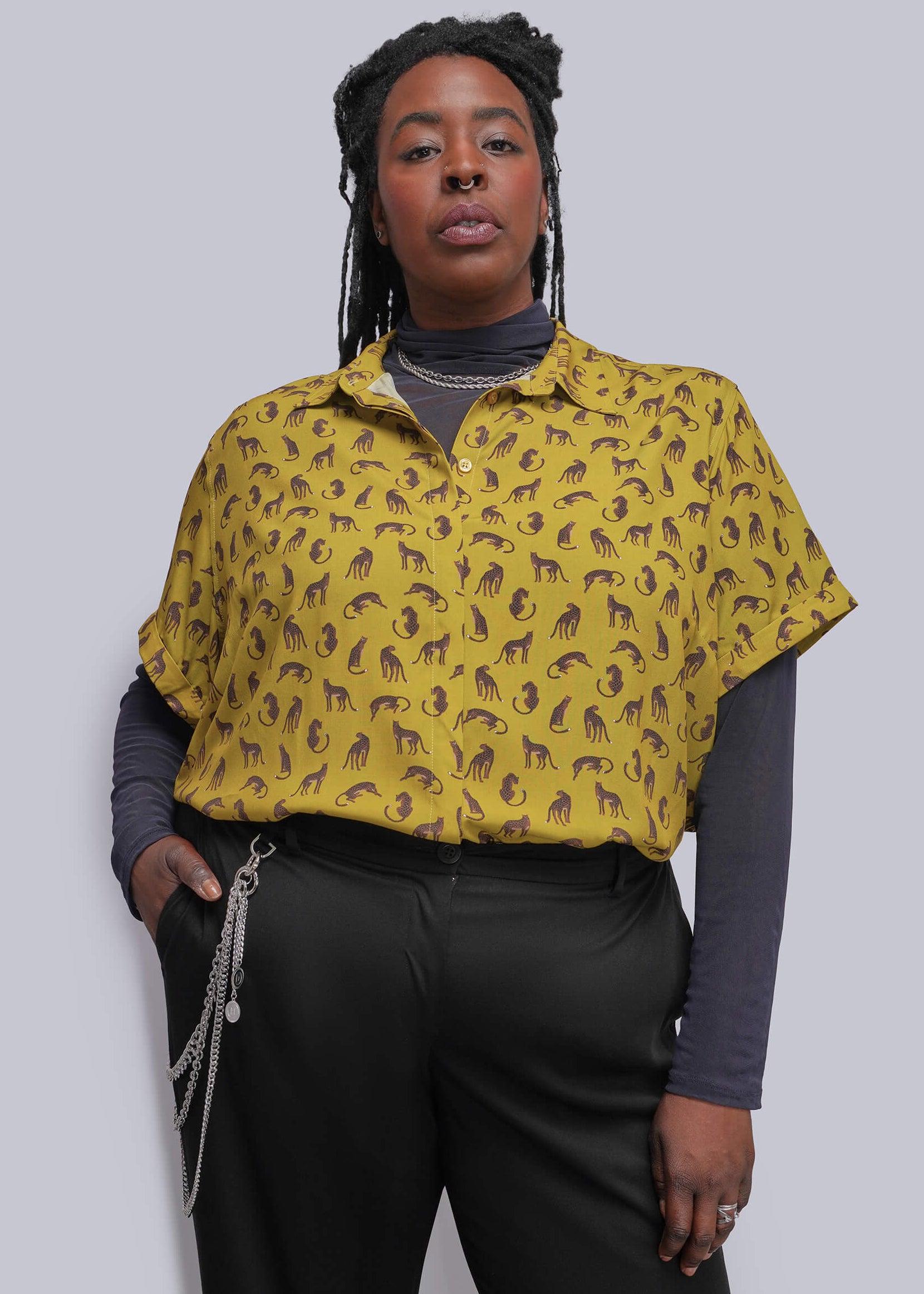 1X=A model wearing a yellow short-sleeve button-up shirt with a brown feline pattern, layered over a dark gray turtleneck and black trousers, against a solid gray background.