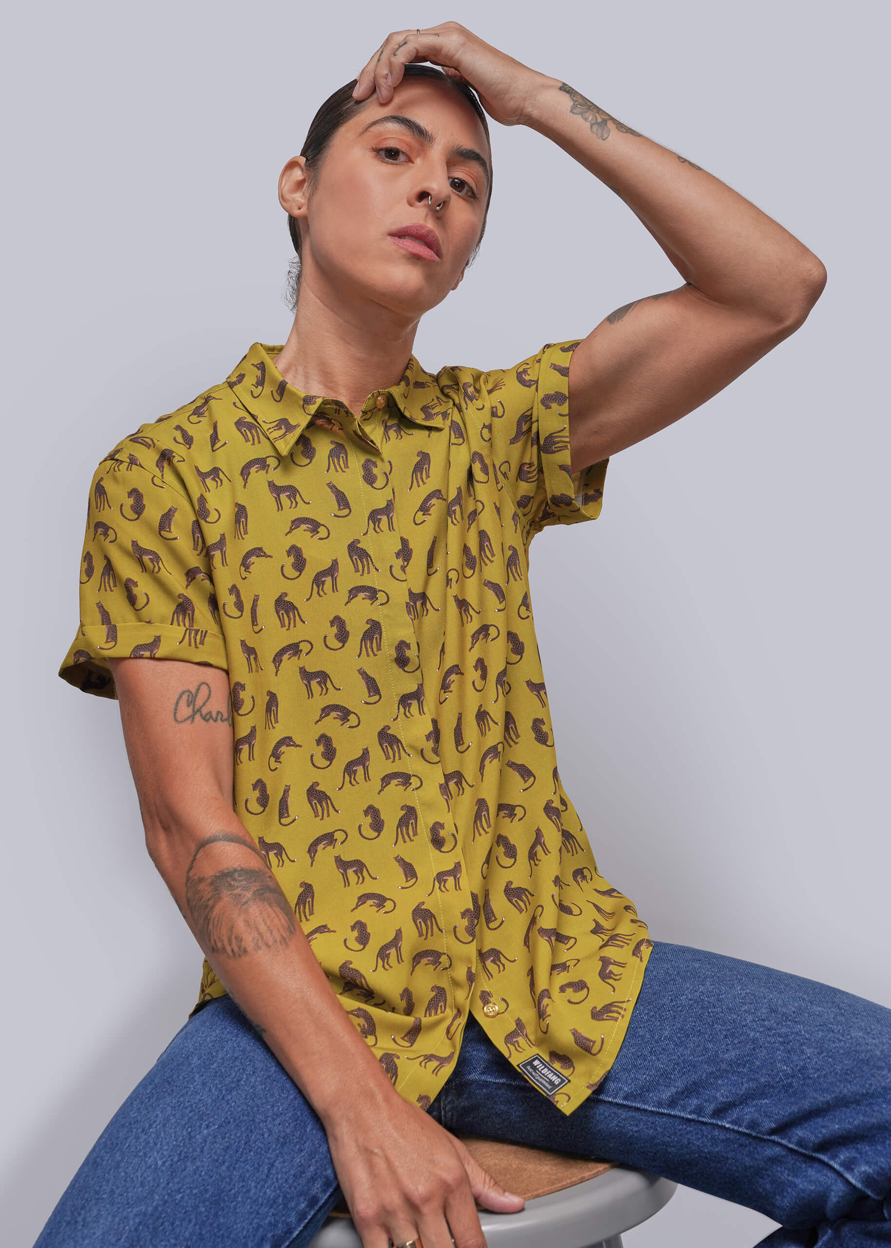 S=A model wearing a yellow short-sleeve button-up shirt with a brown animal print, paired with blue jeans, sitting on a stool against a light gray background.