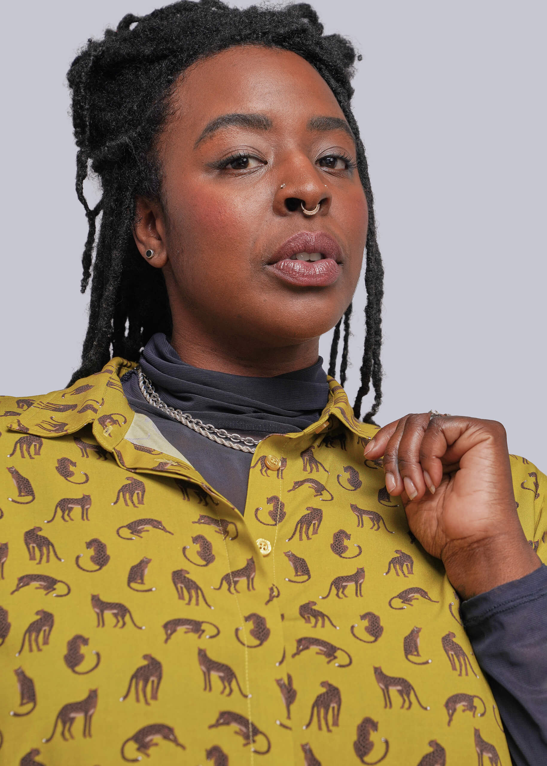 1X=A close-up shot of a person wearing a mustard yellow button-up shirt with a repeating dark brown cat silhouette pattern, layered over a dark gray turtleneck. The background is a solid light gray studio backdrop.