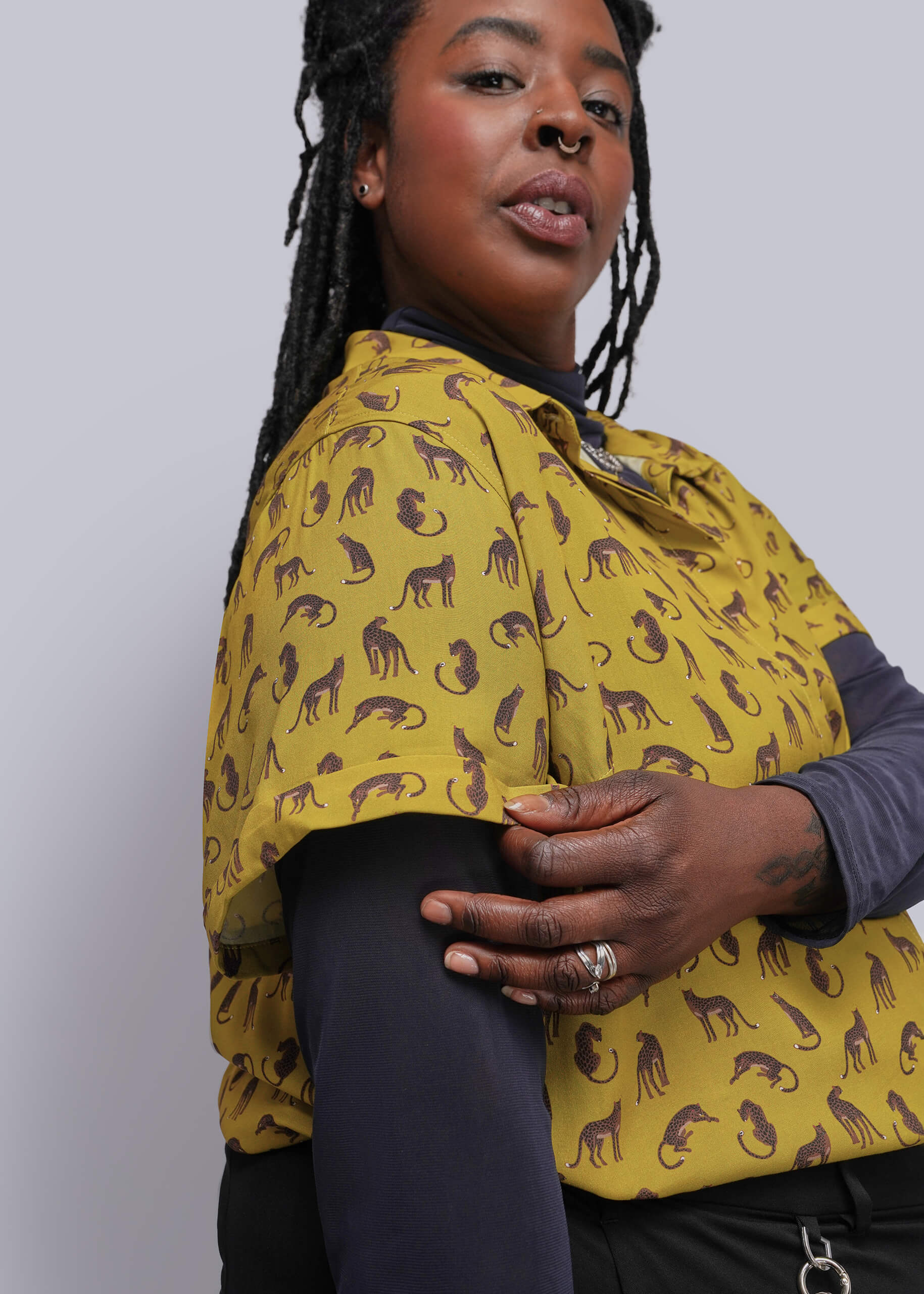 1X=A woman wearing a mustard yellow short-sleeve button-up shirt with a brown animal silhouette print, layered over a dark gray long-sleeve top and black leggings, against a plain light gray background.