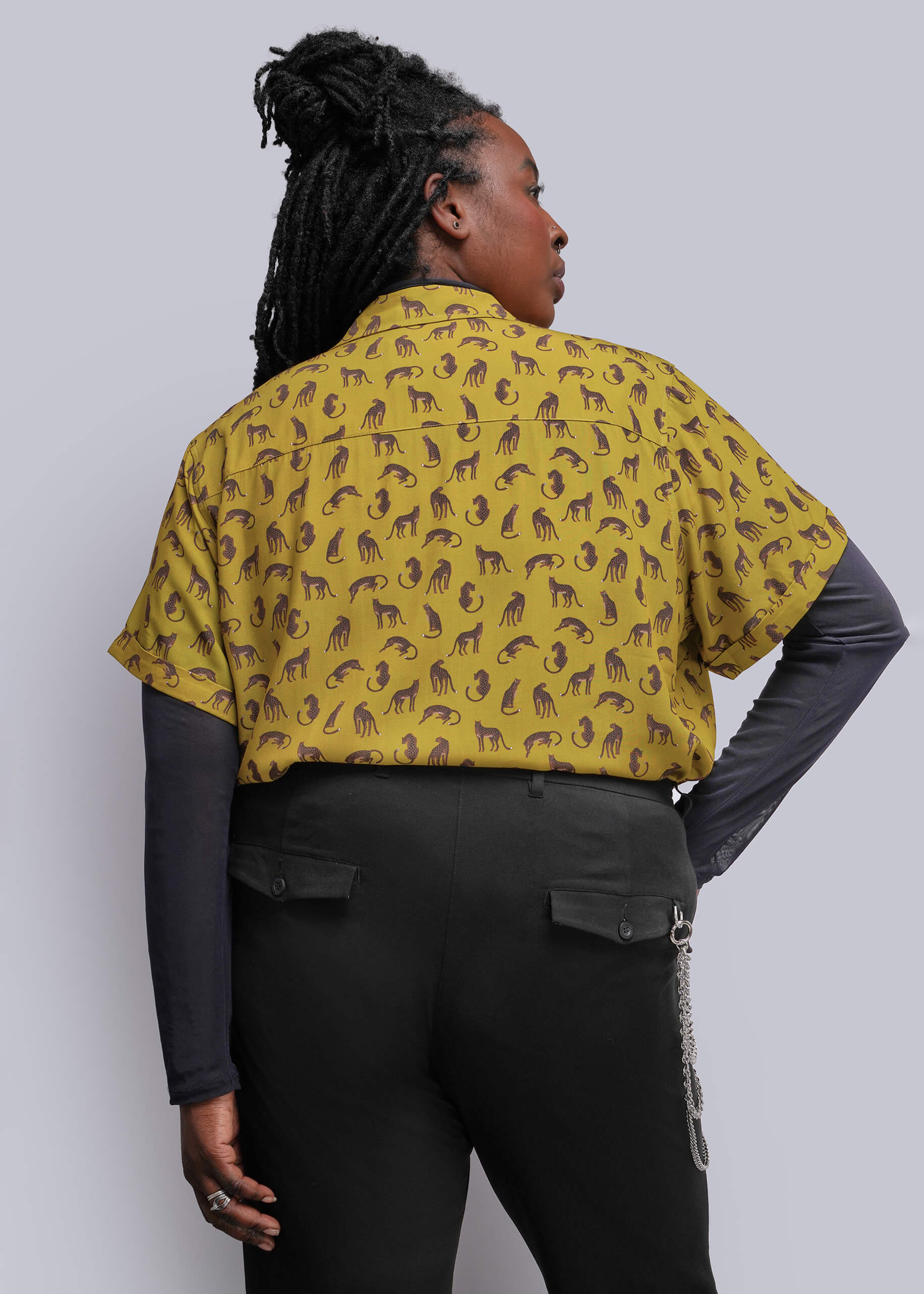 1X=A woman shown from the back wearing a mustard yellow short-sleeve button-up shirt with a brown pattern, layered over a dark gray long-sleeve top and black pants, against a solid light gray background.