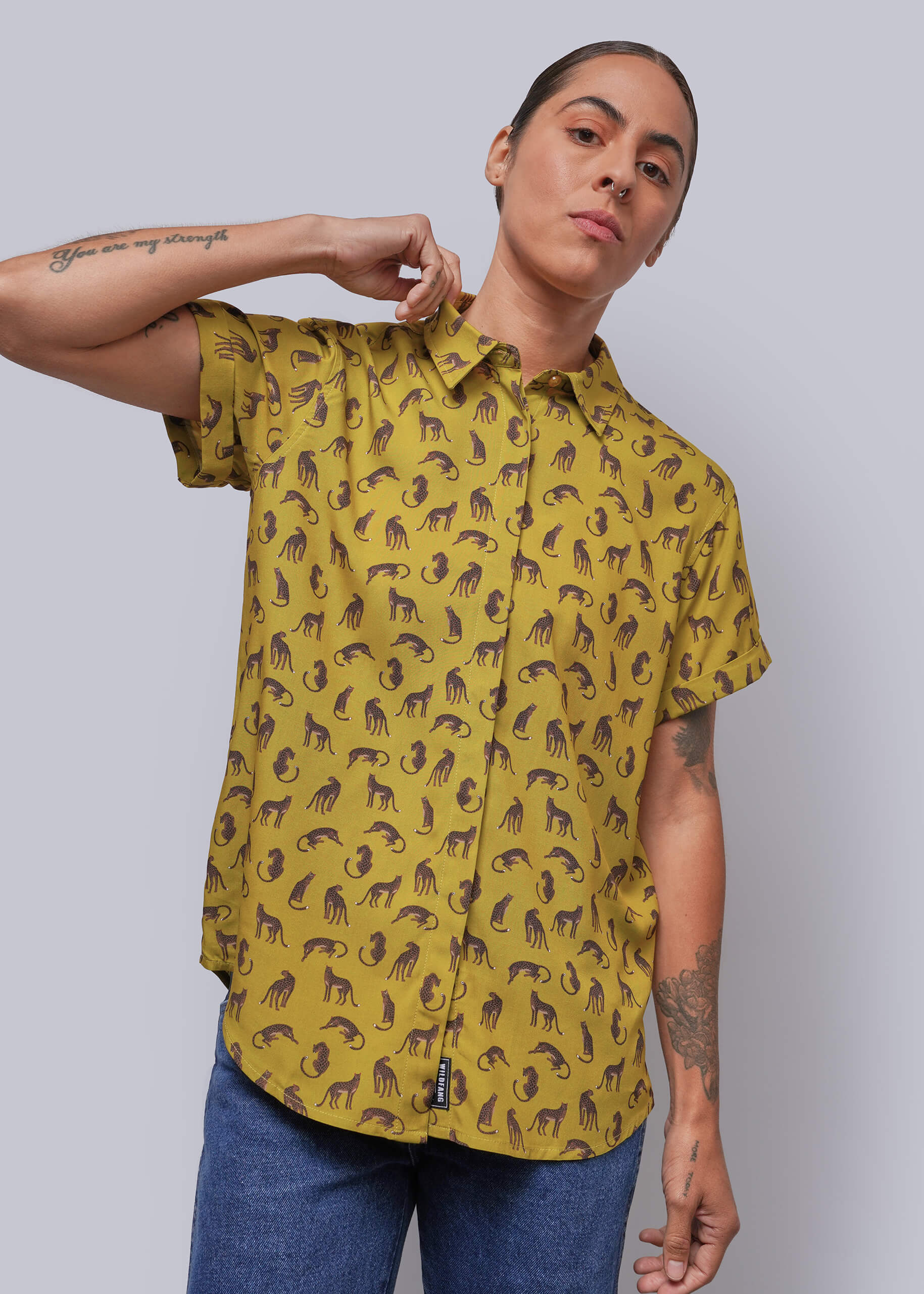 S=A person wearing a mustard yellow short-sleeve button-up shirt with a repeating animal print, paired with blue jeans, against a solid gray background.
