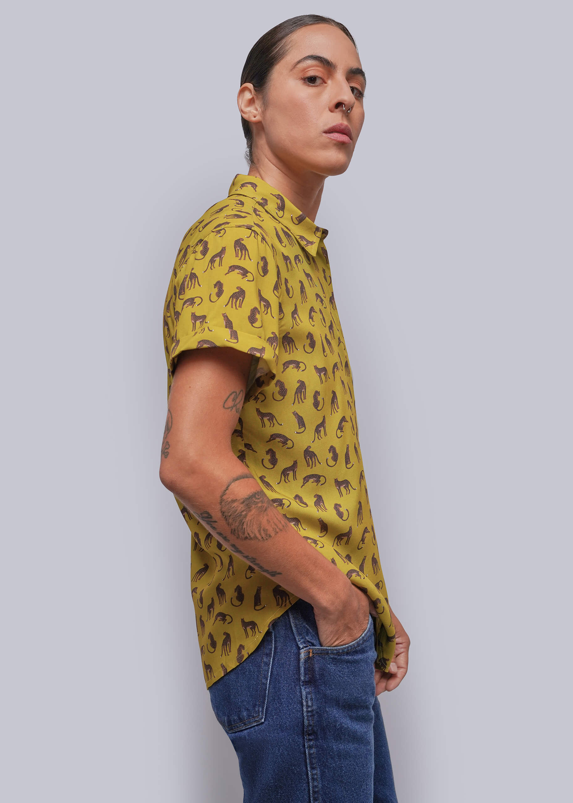 S=A person wearing a yellow short-sleeve button-up shirt with a brown feline pattern, paired with blue jeans, photographed against a solid gray background.