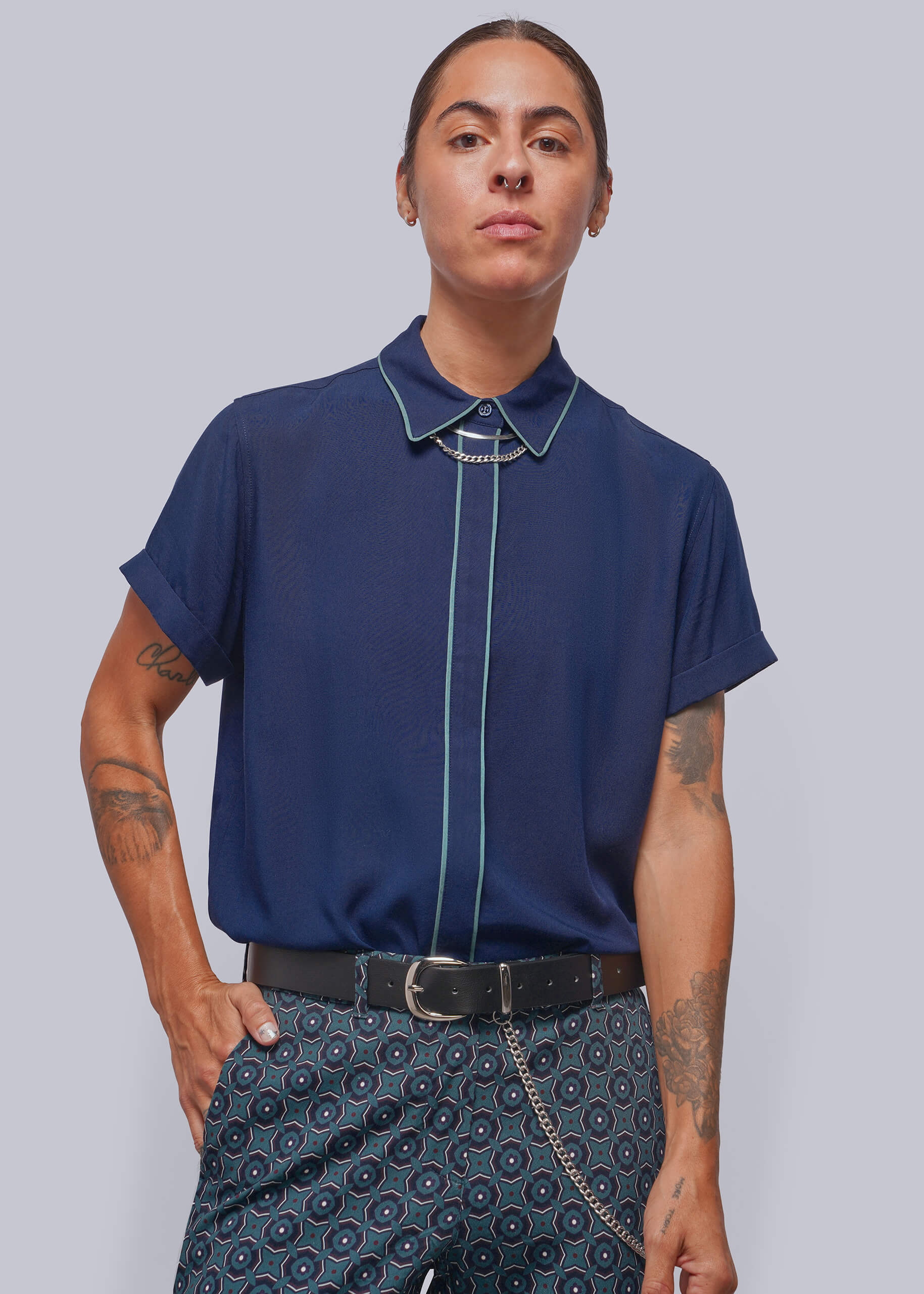 A model wearing a navy blue short-sleeve button-up shirt with light blue piping details on the collar, front placket, and cuffs, paired with patterned trousers, against a solid gray background.