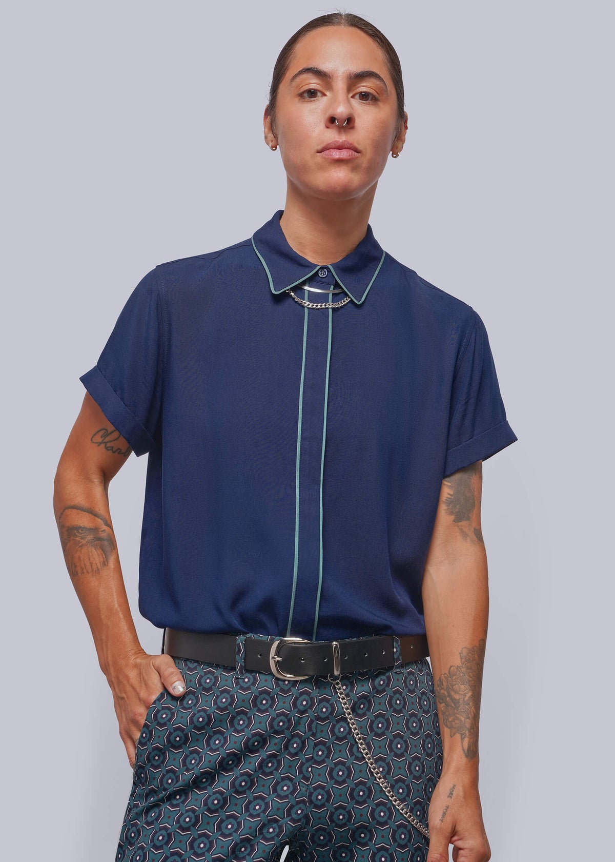 S=A model wearing a navy blue short-sleeve button-up shirt with light blue piping details on the collar, front placket, and cuffs, paired with patterned trousers, against a solid gray background.
