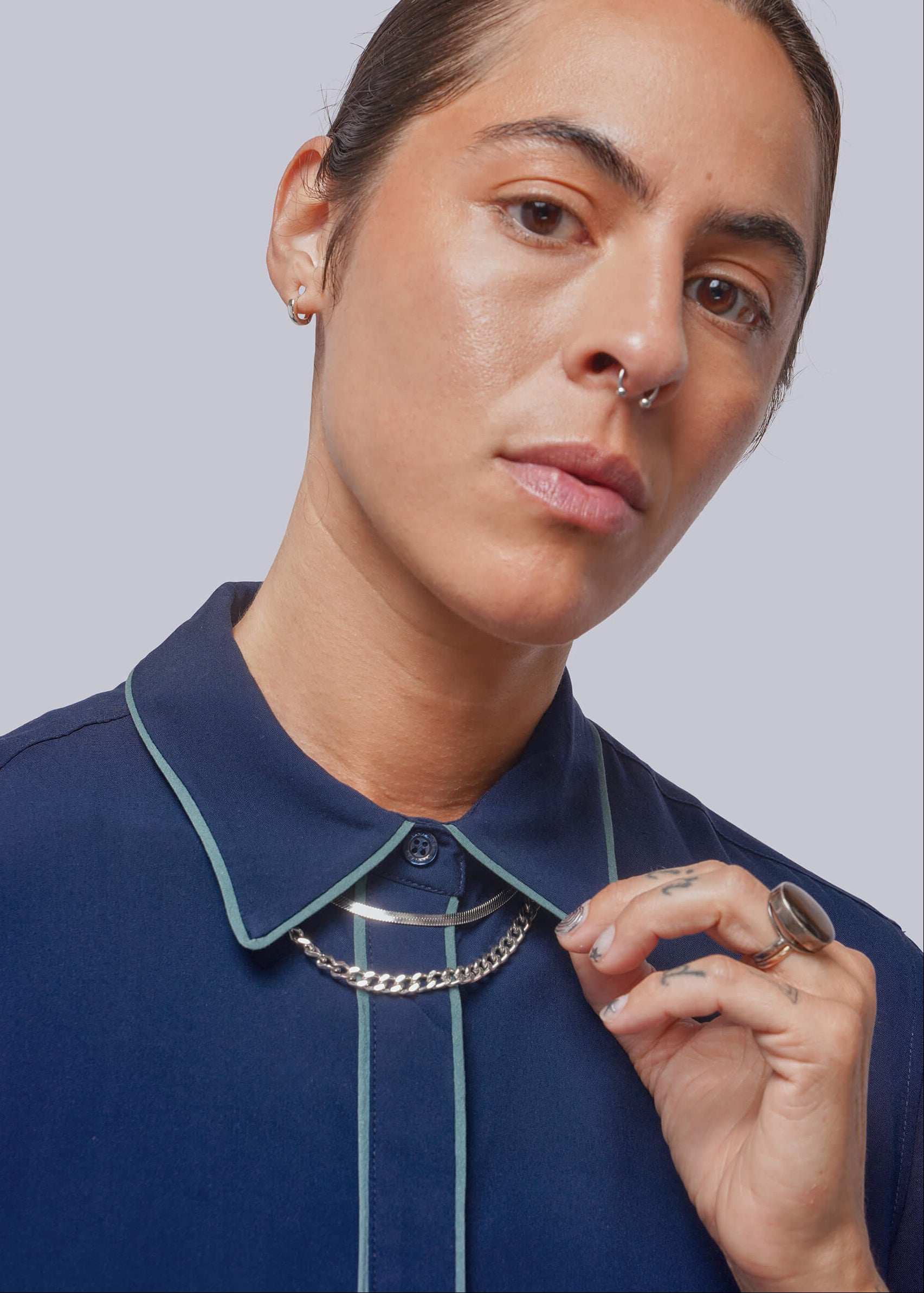 A close-up of a model wearing a navy blue collared shirt with light green piping and a chunky silver chain necklace, set against a solid gray background.