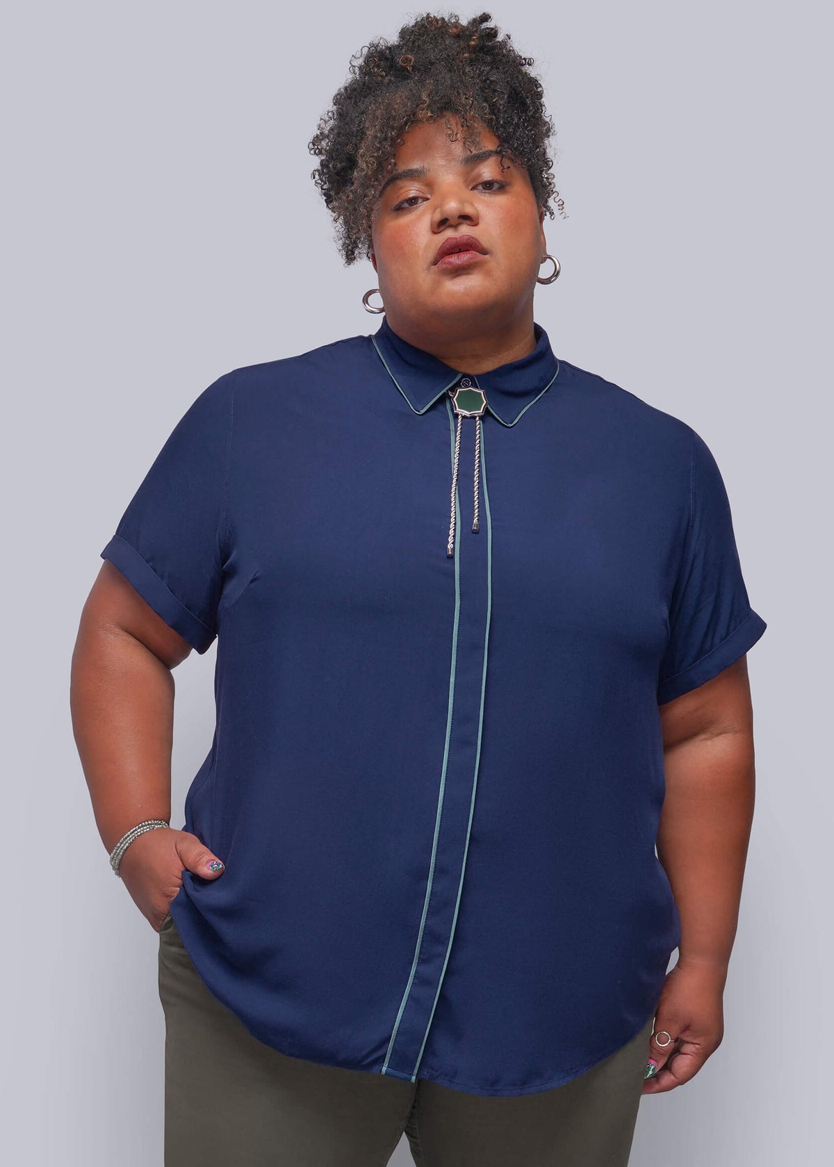 1X=A plus-size model wearing a navy blue short-sleeve button-up blouse with contrasting light blue piping and a decorative tie-neck detail, paired with olive green pants, against a solid gray studio background.