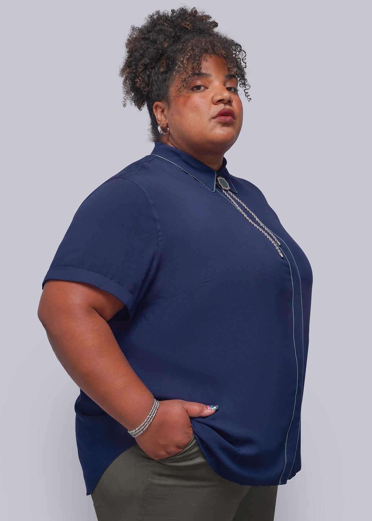 1X=A woman modeling a navy blue short-sleeve button-up shirt with a silver zipper detail at the collar, paired with olive green pants, against a solid light gray background.