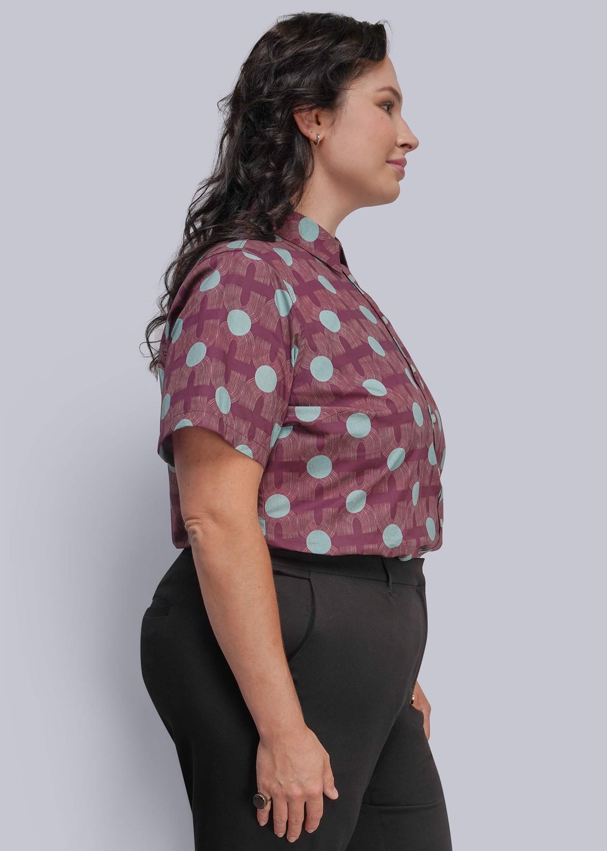 1X=A woman in side profile wearing a short-sleeve button-up shirt with a purple and light blue geometric pattern, paired with black pants, against a solid light gray background.