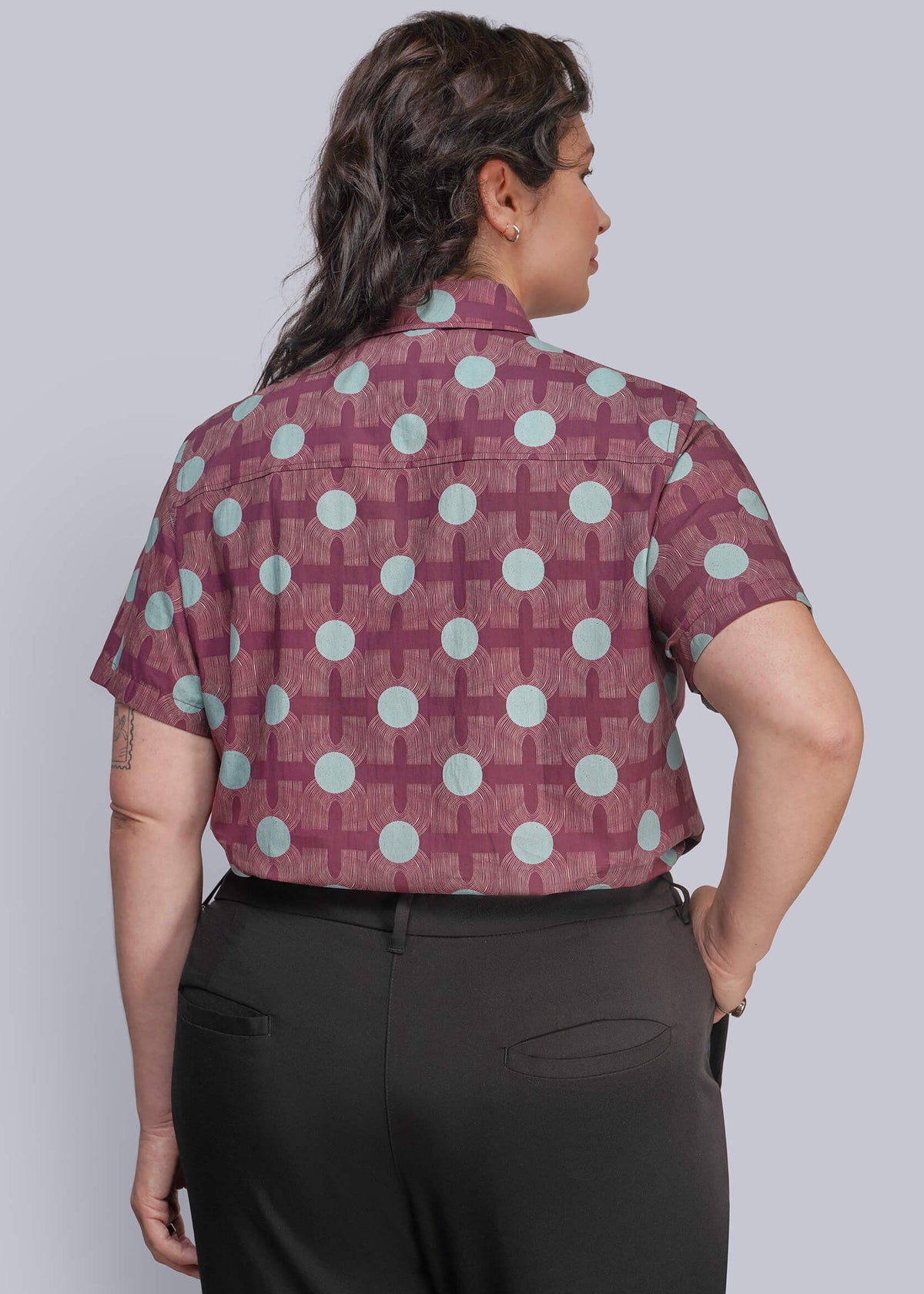 1X=A woman shown from the back wearing a short-sleeve button-up shirt with a burgundy and light blue geometric pattern, paired with black trousers, against a solid gray studio background.