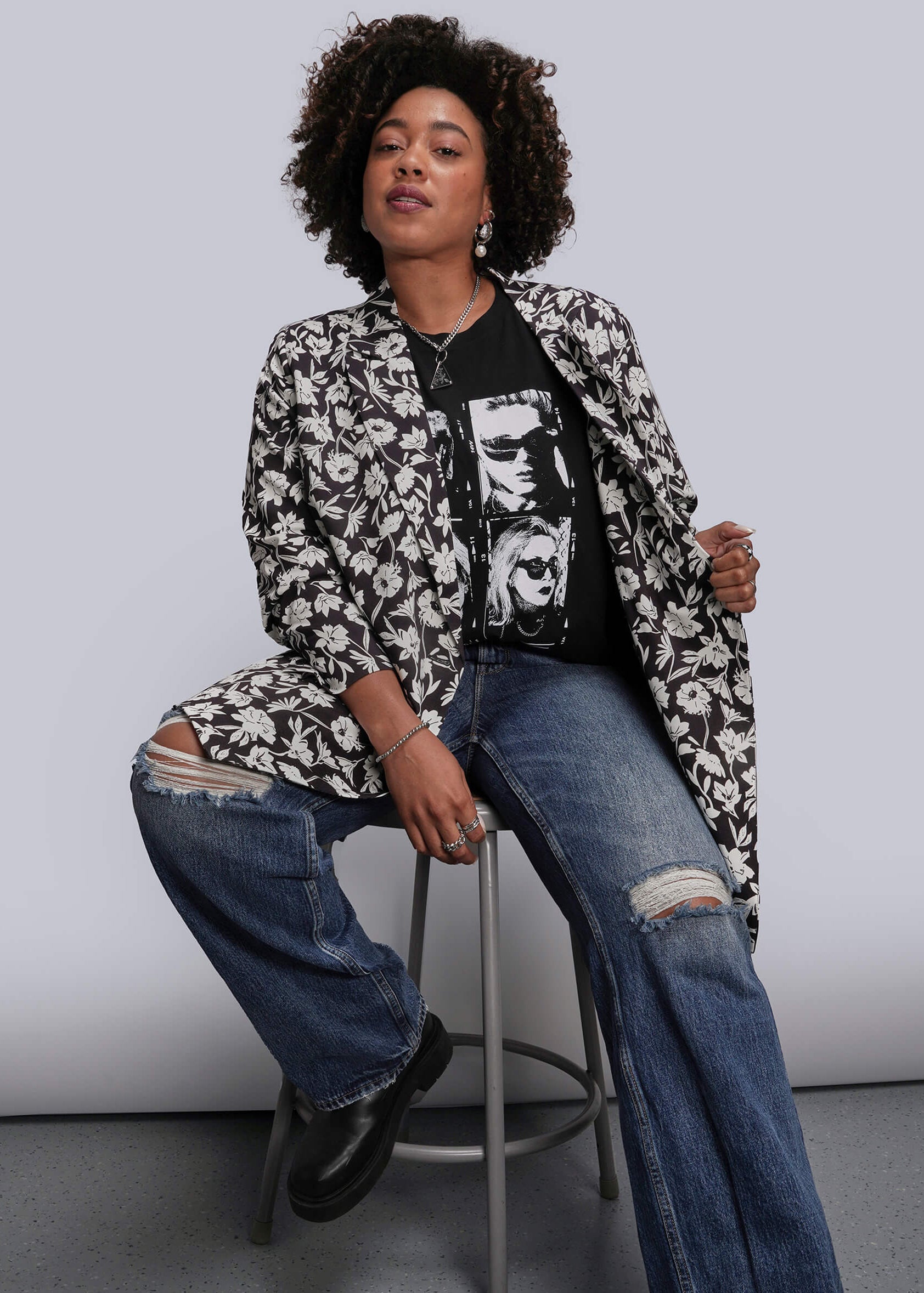 1X=Person sitting on a stool wearing a floral blazer, graphic t-shirt, and jeans against a plain background