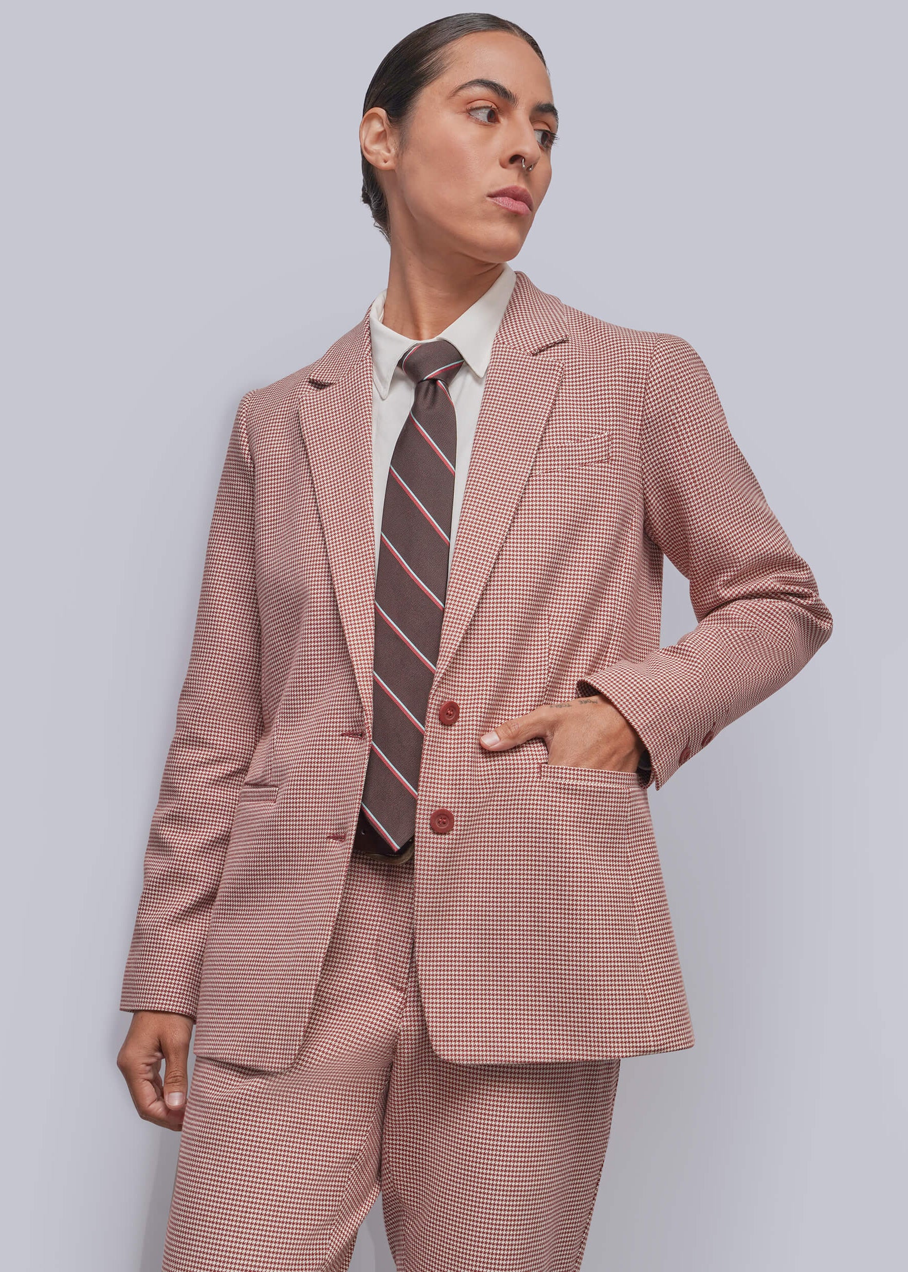 S=A model wearing a cinnamon and white micro-check suit, including a blazer and trousers, styled with a white collared shirt and a brown and pink striped tie, against a solid gray background.