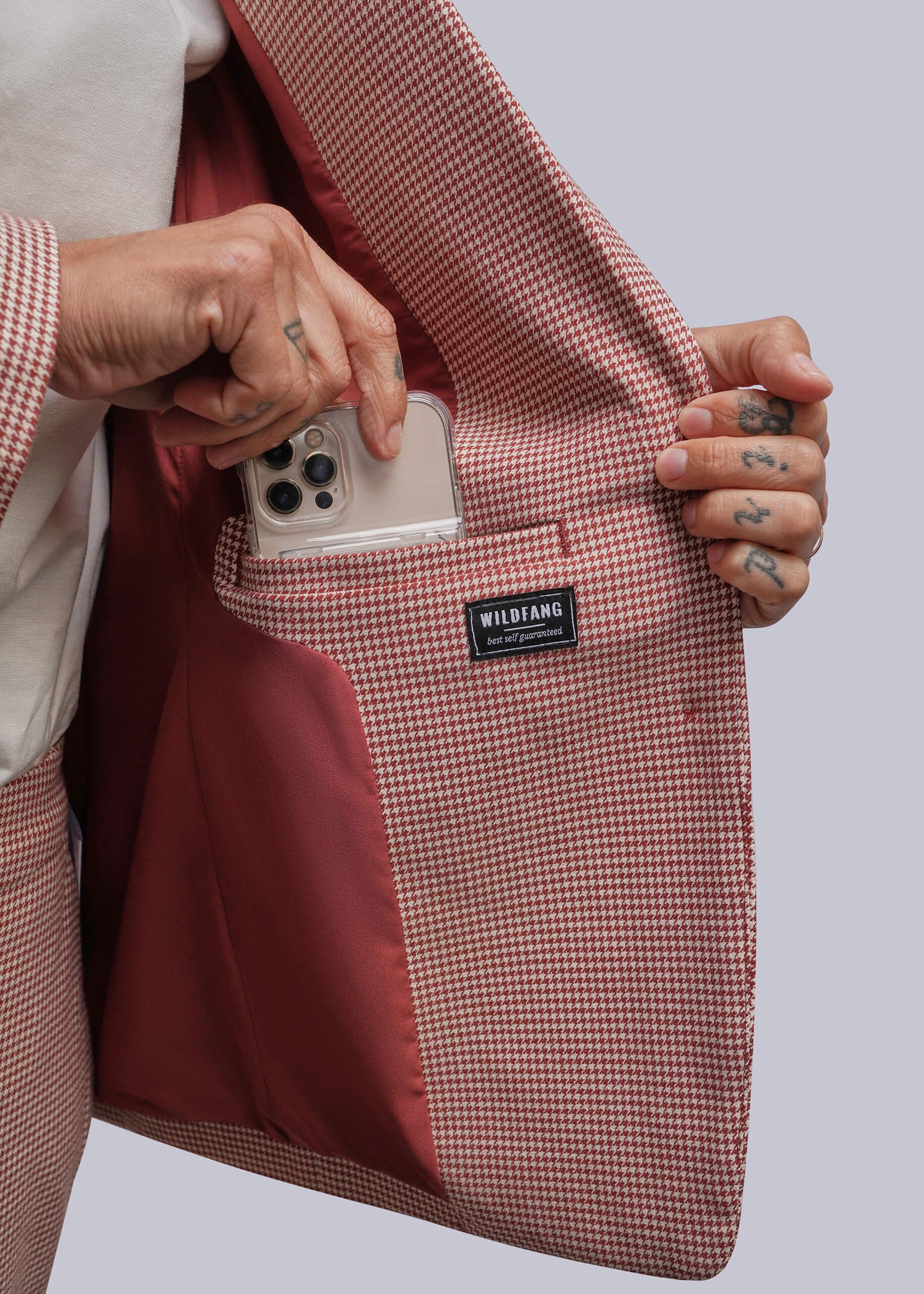 A close-up of a person holding open a dusty red and white micro-check blazer to show a phone pocket with a red lining. A black brand label reading WILDFANG is visible on the pocket.
