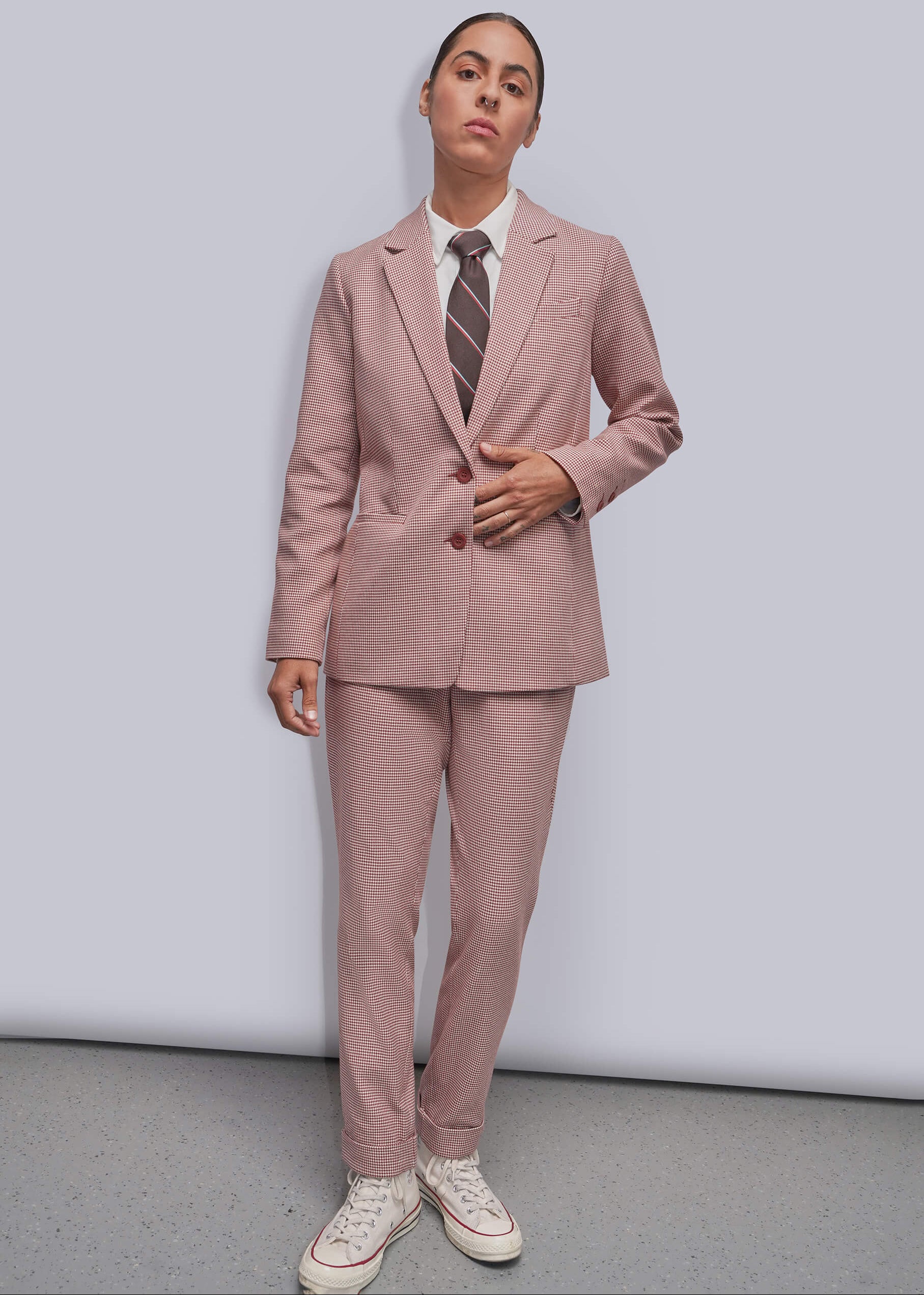 S=A model wearing a dusty red two-piece suit with a white collared shirt and a striped tie, paired with white sneakers, standing against a light gray studio background.