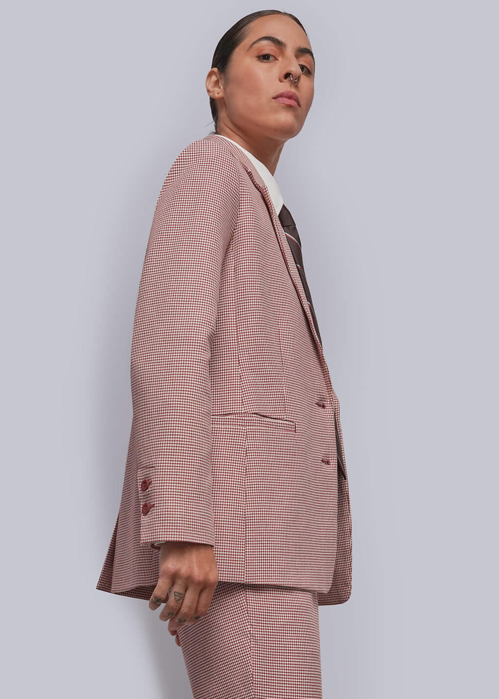 S=A model is shown in profile wearing a dusty red two-piece suit with a white collared shirt and a dark patterned tie, paired with white canvas sneakers, against a light gray studio background.